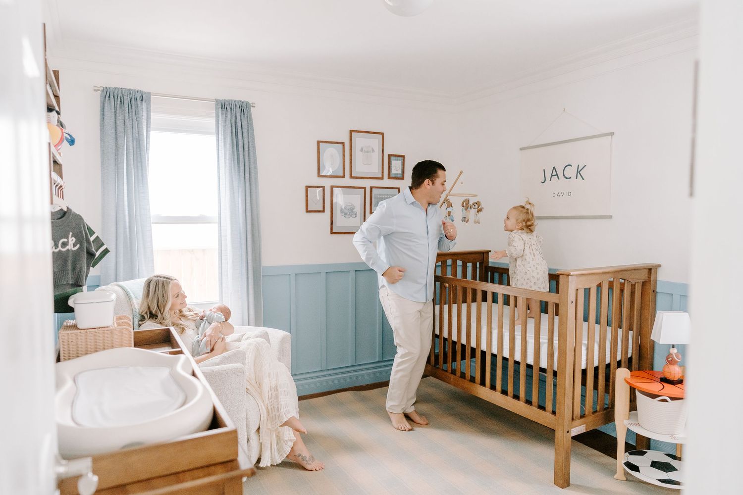 Father standing in a bright nursery with toddler and newborn baby in wooden crib.
