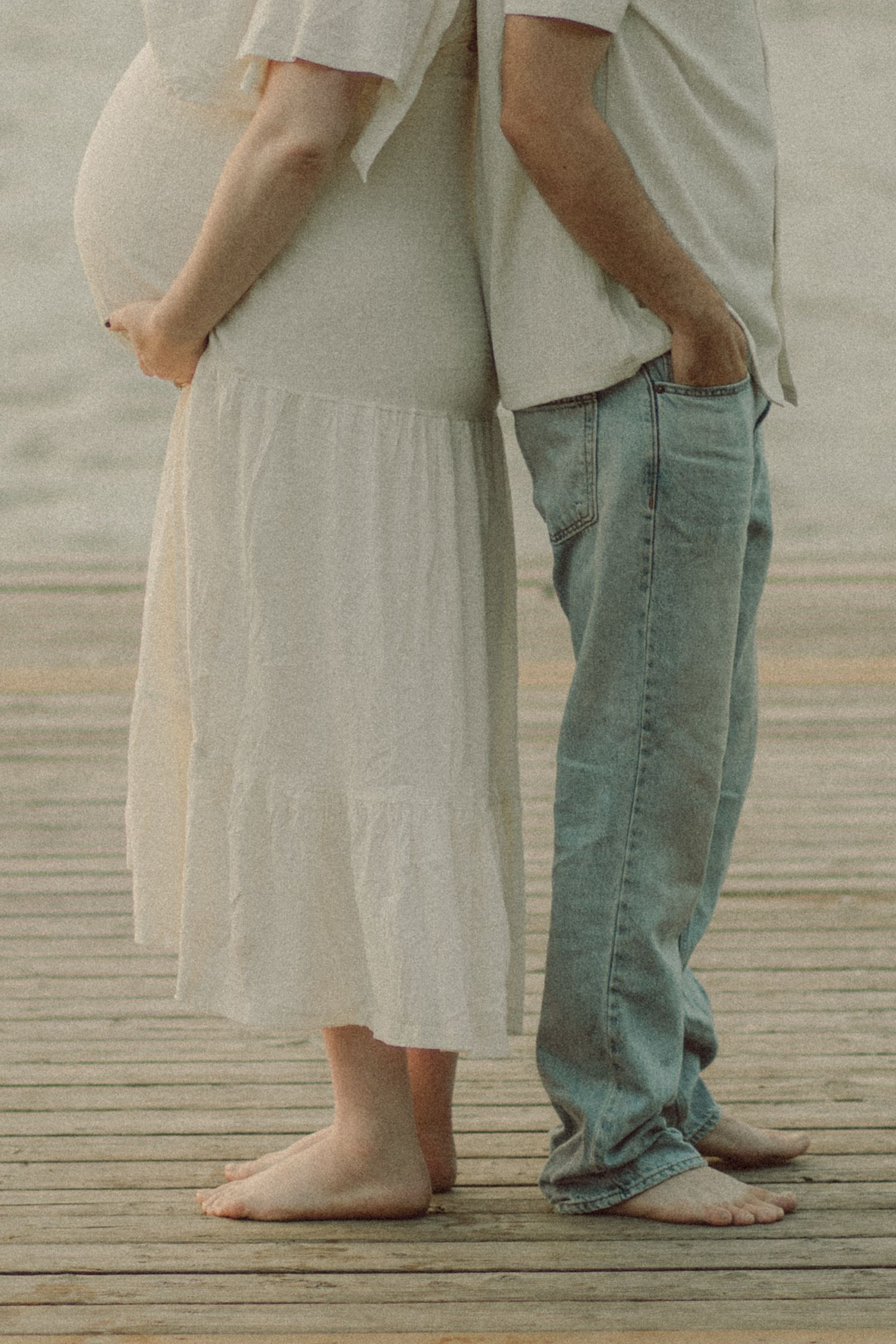 Pregnant couple standing barefoot on wooden dock, woman in white dress showing baby bump, man in jeans.