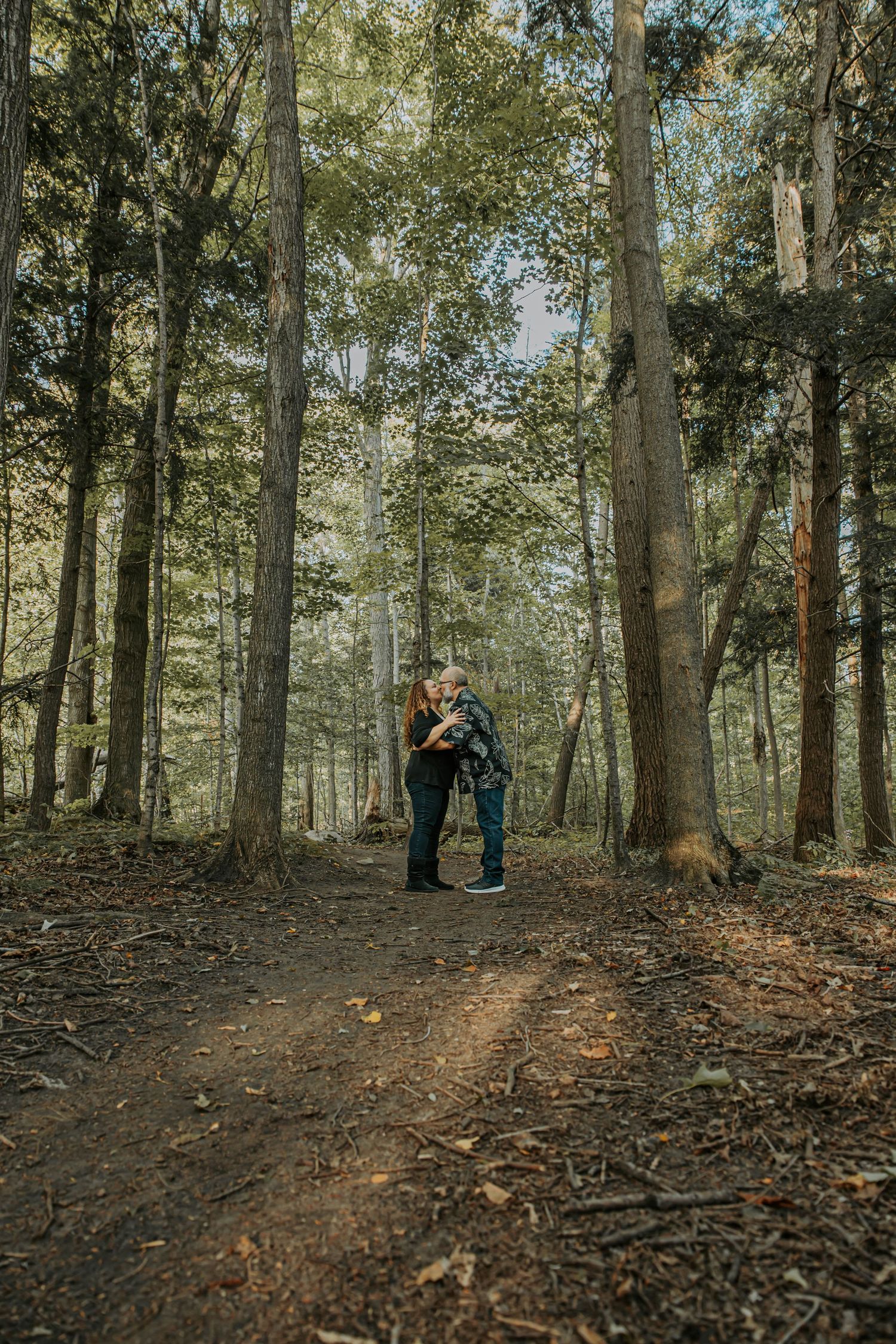 Two figures stand in a atmospheric wooded area during autumn with fallen leaves scattered on the forest floor.