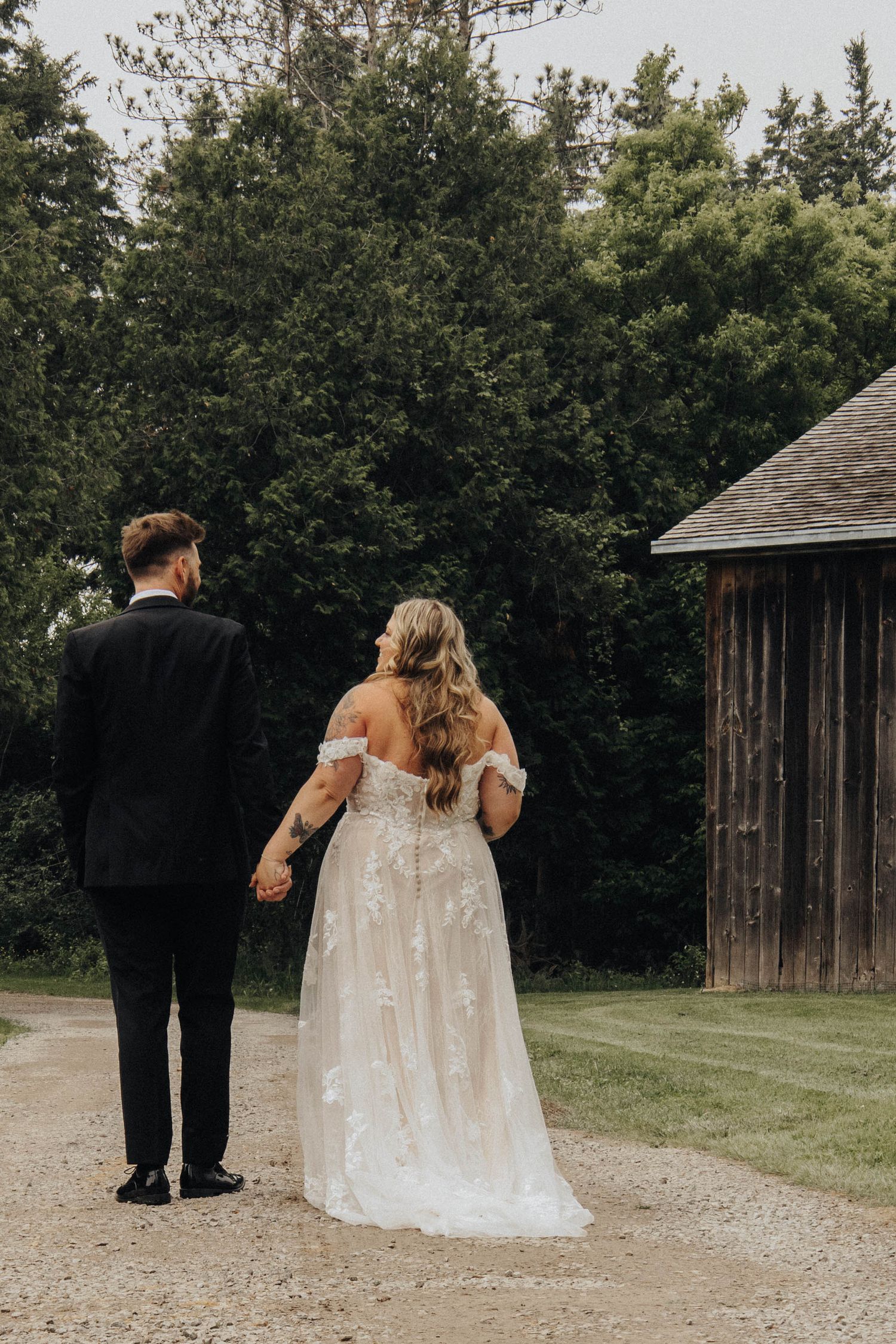 Wedding couple walking away together on gravel path with rustic barn in background.