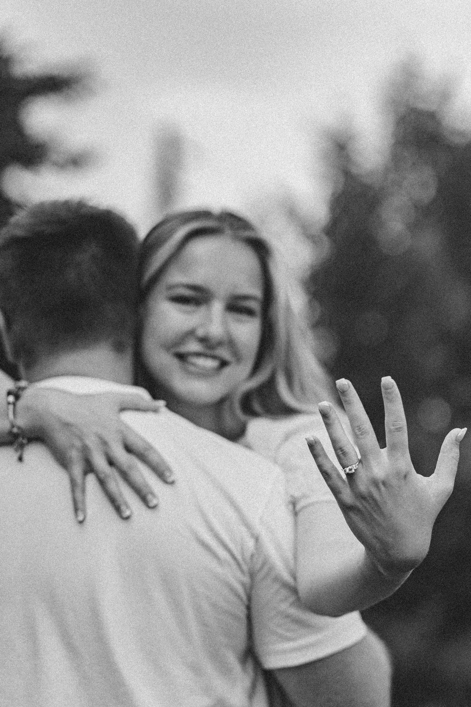 A black and white photo showing hands with engagement rings raised in celebration against a blurred outdoor background.