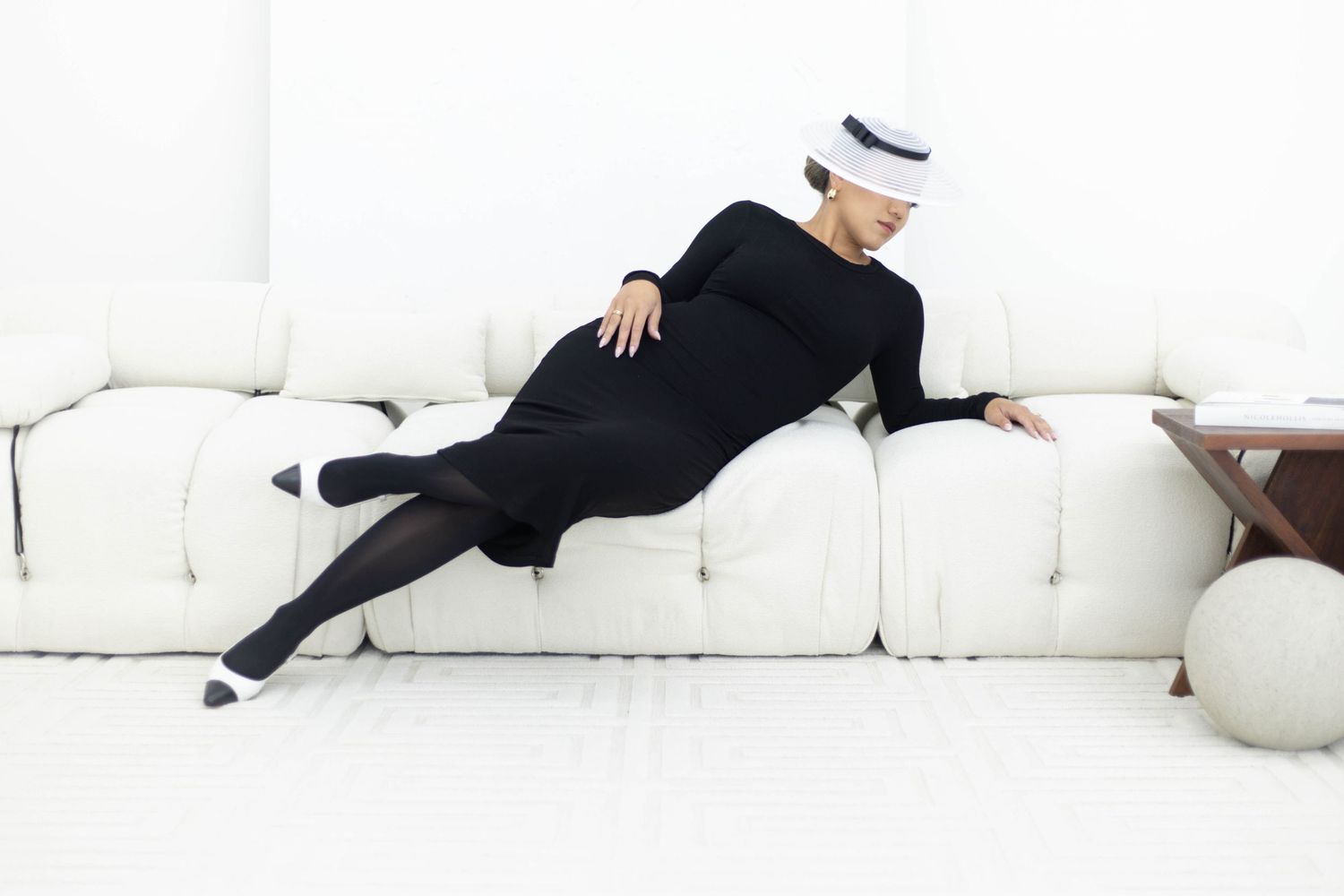 Someone in an all black outfit and white hat lounging on a white couch with artistic pose.