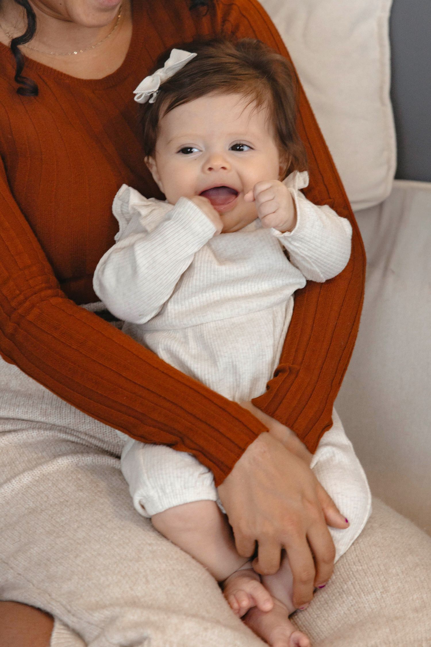 Smiling infant in white clothing lies on a textured surface while supported by hands in a brown sweater.