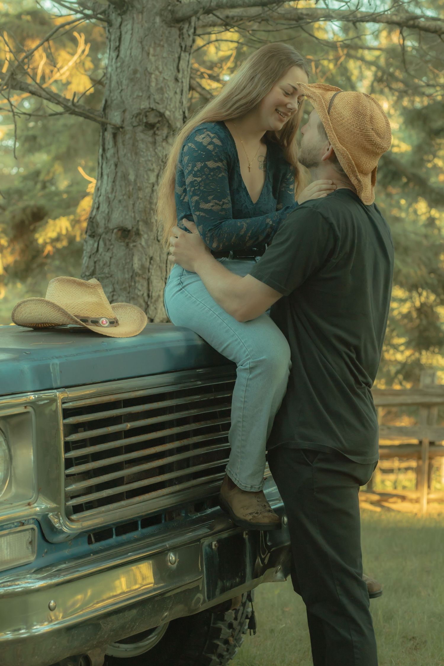 Romantic couple near a vintage truck, woman sitting on hood, man in straw hat gazing up at her.