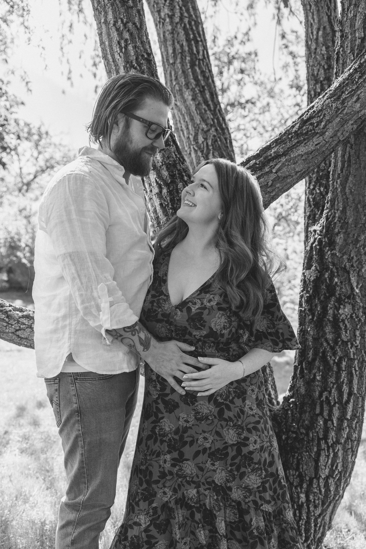 Black and white photo of a pregnant couple embracing tenderly by a tree outdoors.