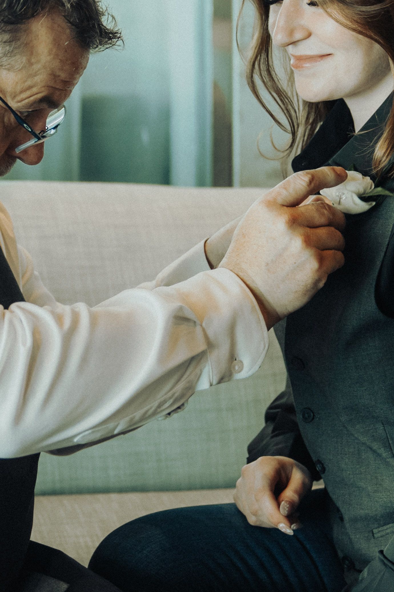 An older man pins a white boutonniere on a smiling young woman in a grey suit at a wedding.