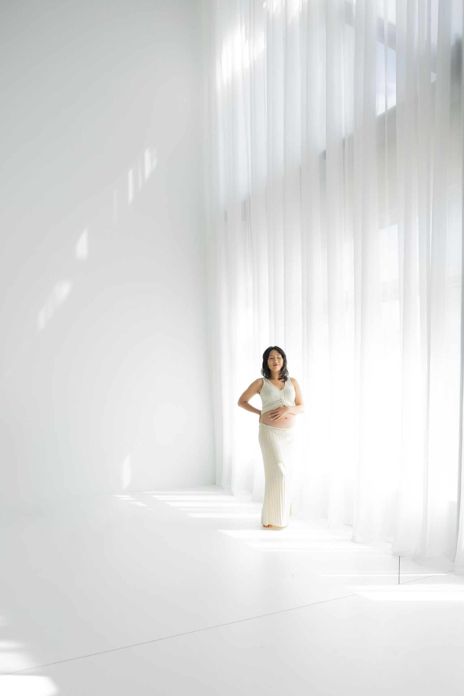 Pregnant woman in white dress standing in bright white room with sheer curtains, cradling her baby bump.