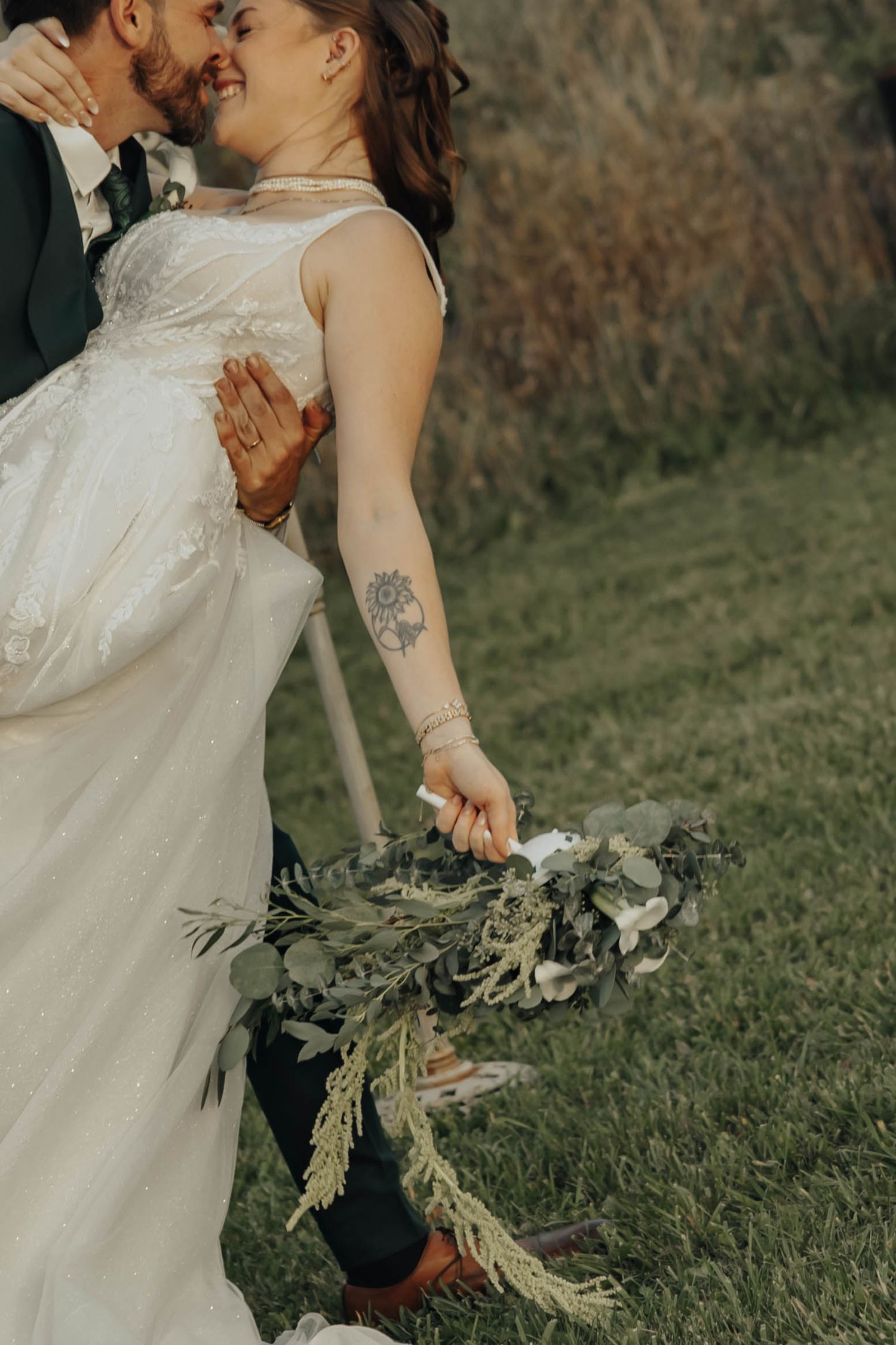 A couple shares an intimate moment outdoors while holding a rustic bouquet with greenery.