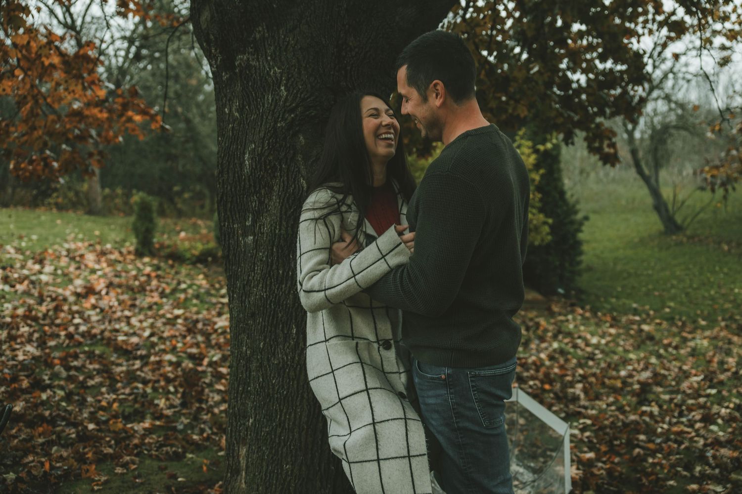 A couple shares an intimate moment against a tree in an autumn park with fallen leaves scattered on the ground.
