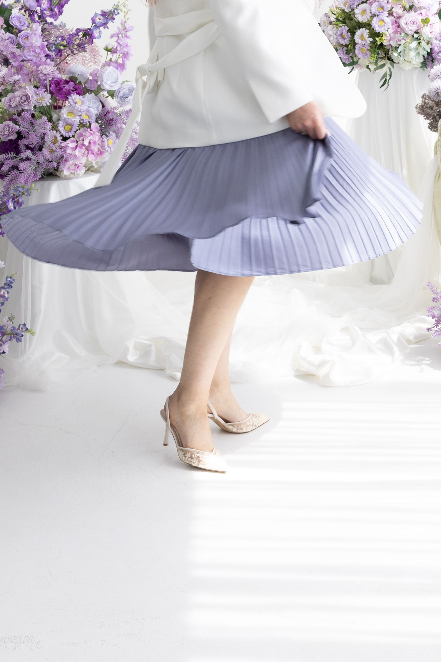 Woman in white jacket twirling blue pleated midi skirt wearing nude heels with purple floral backdrop.