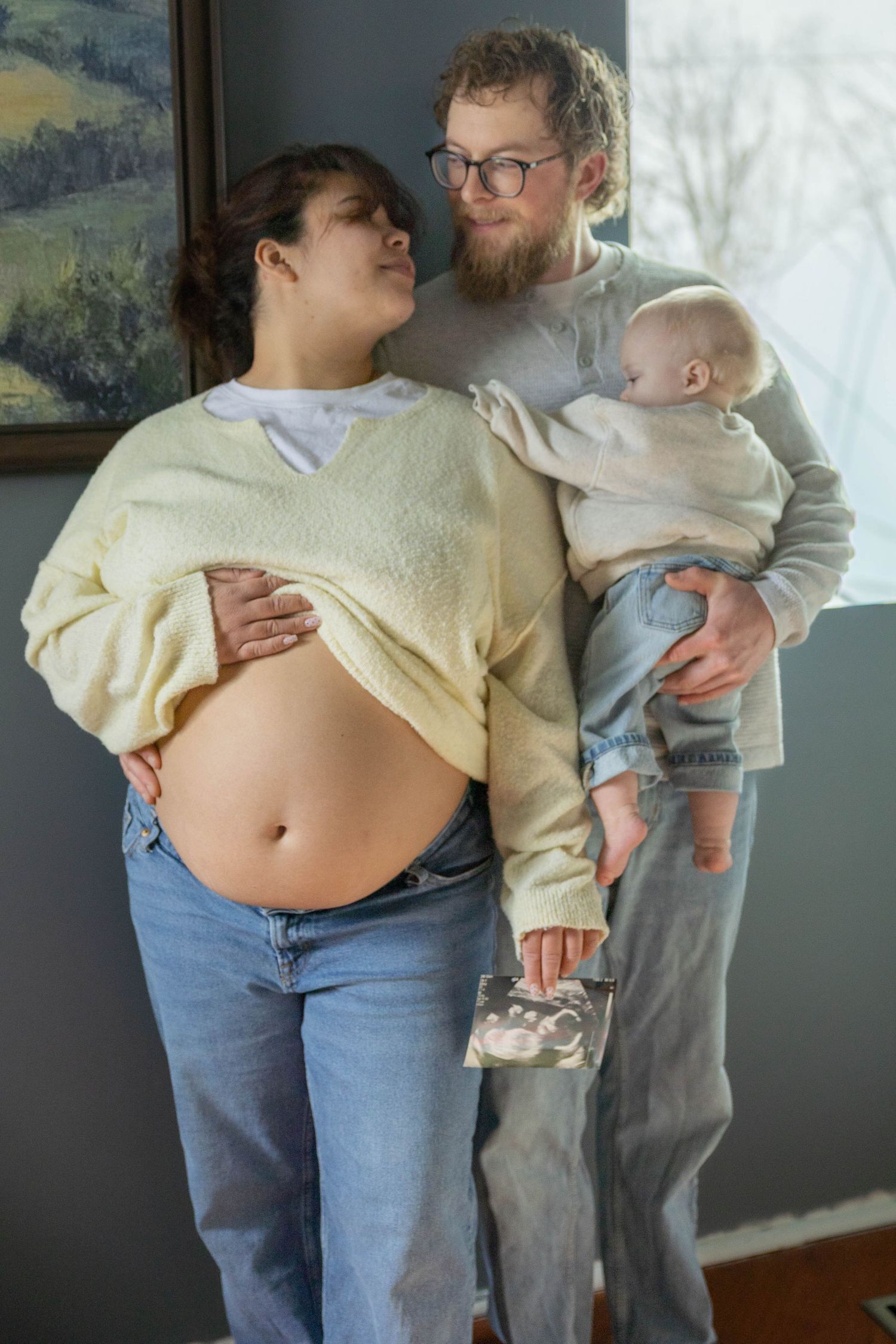 Family stands together showing pregnant belly while wearing casual clothes in front of wall art.