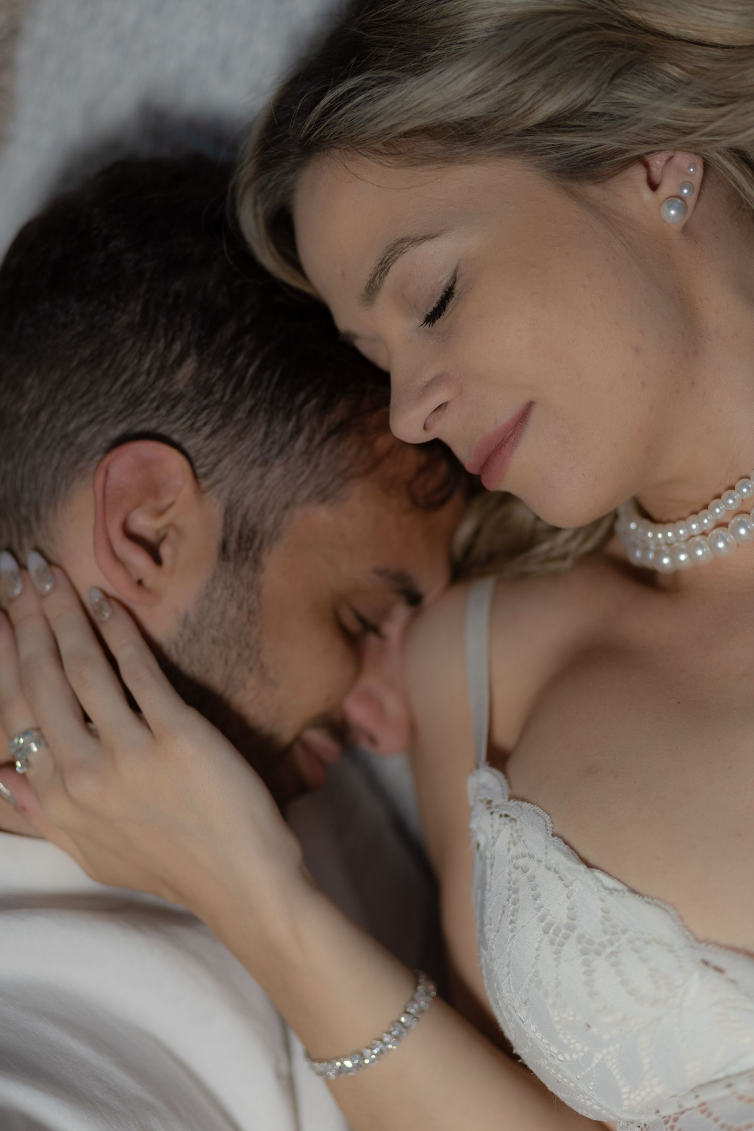 Romantic couple in an intimate moment, woman wearing pearl jewelry and lace lingerie, man in white shirt.