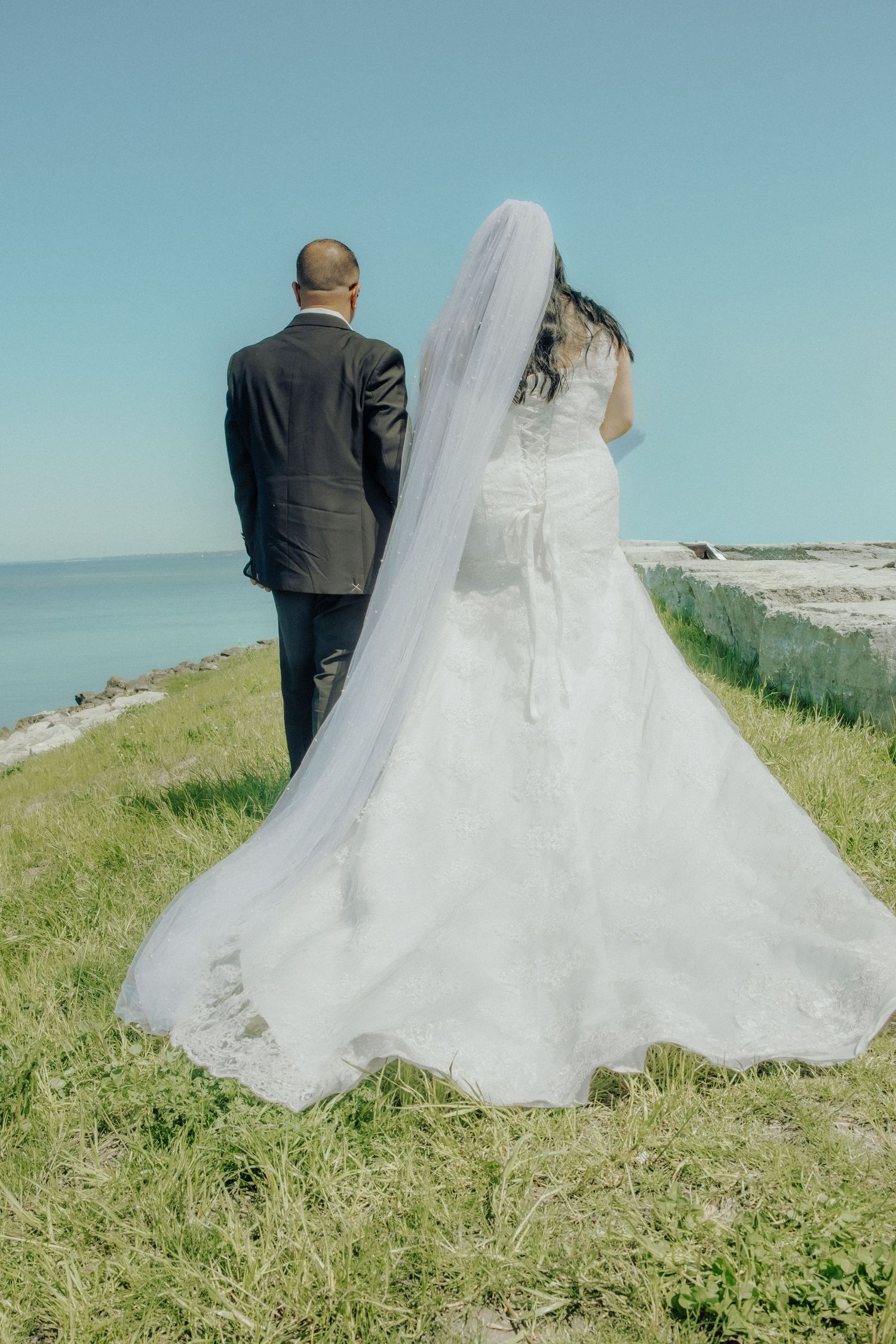 Bride and groom standing together facing the ocean, bride wearing a white lace gown with long veil on a sunny day.