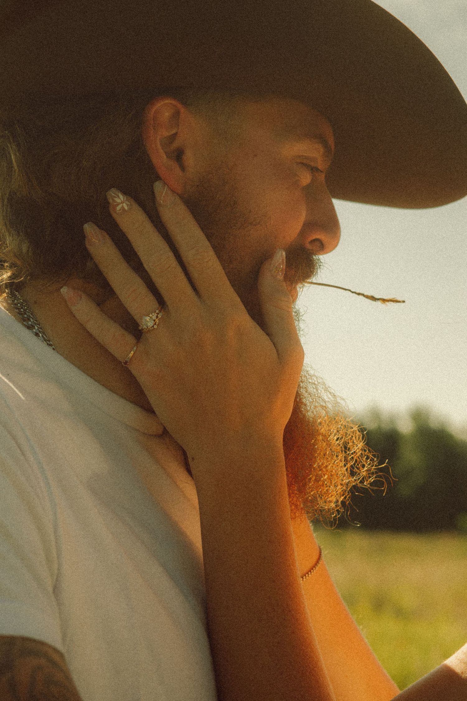 Bearded man in cowboy hat with a woman's hand resting on his face in a sunny field.