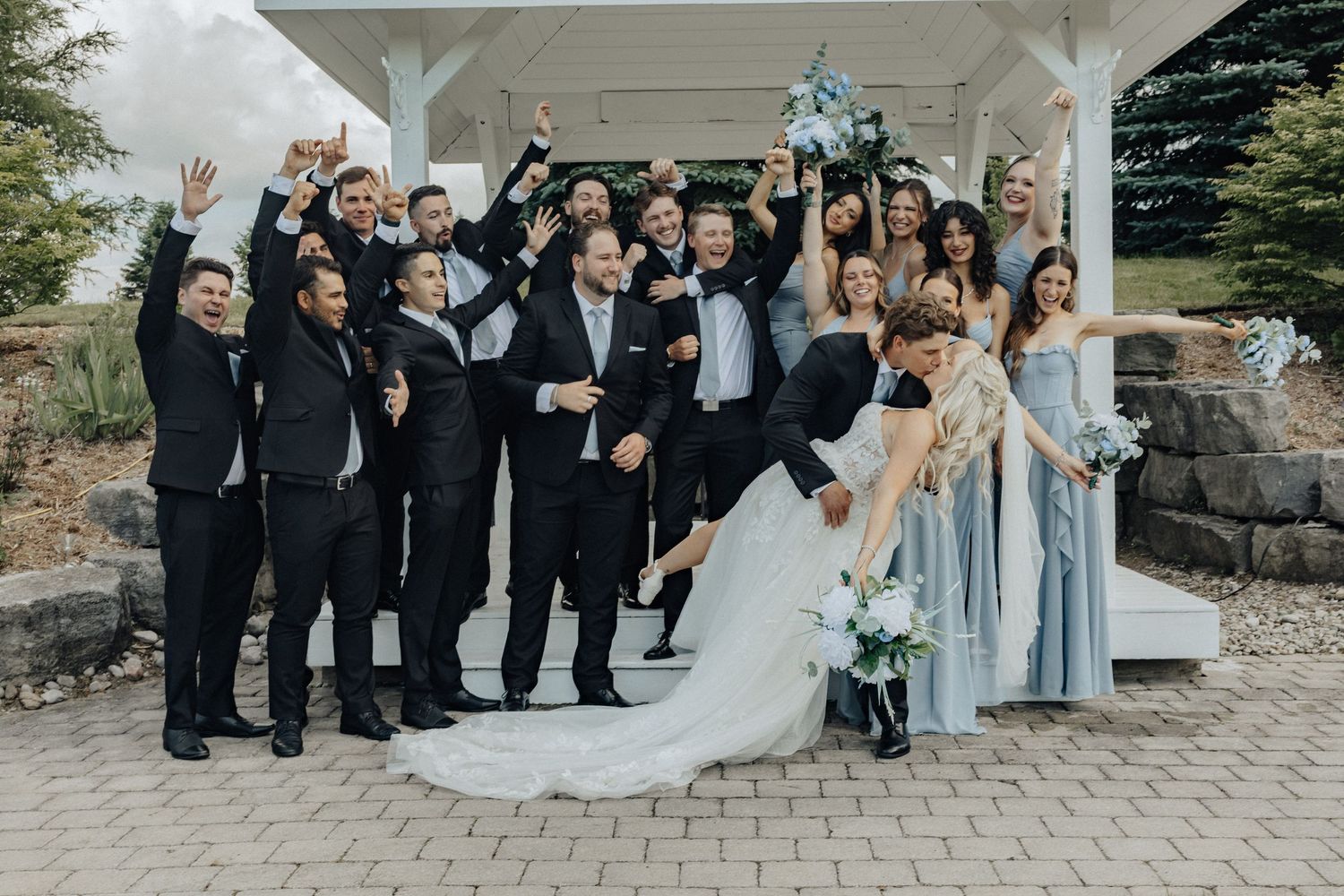 Wedding party celebrating outdoors under a pergola, bride and groom dipping while guests cheer in formal attire.