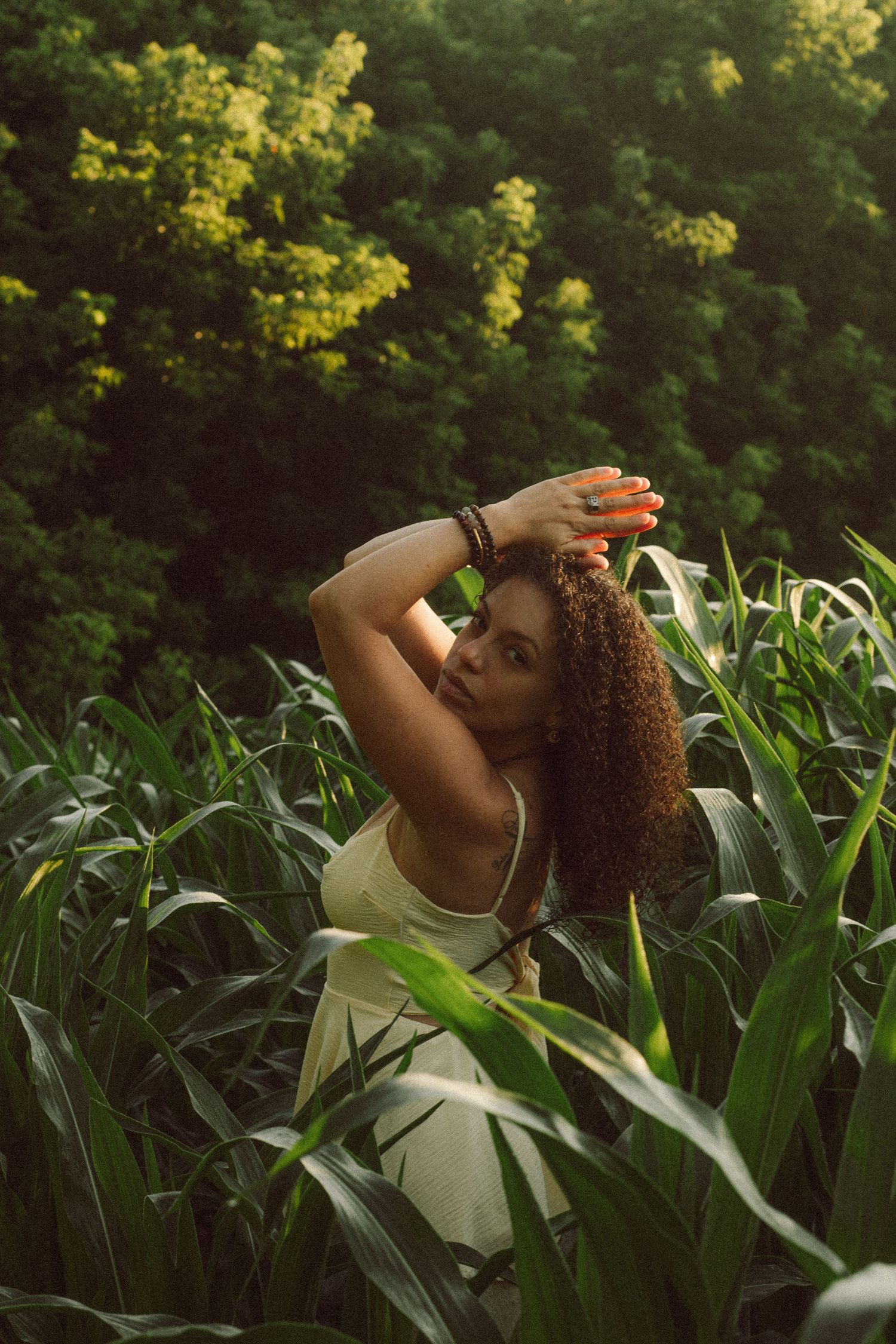 Young woman with curly hair posing gracefully in a lush green cornfield, wearing a light dress with bracelets.
