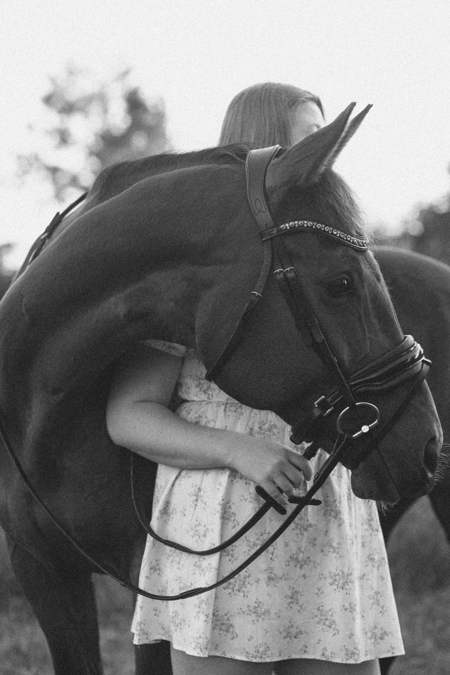 Black and white photo of a young girl in a floral dress hugging a horse wearing a bridle outdoors.