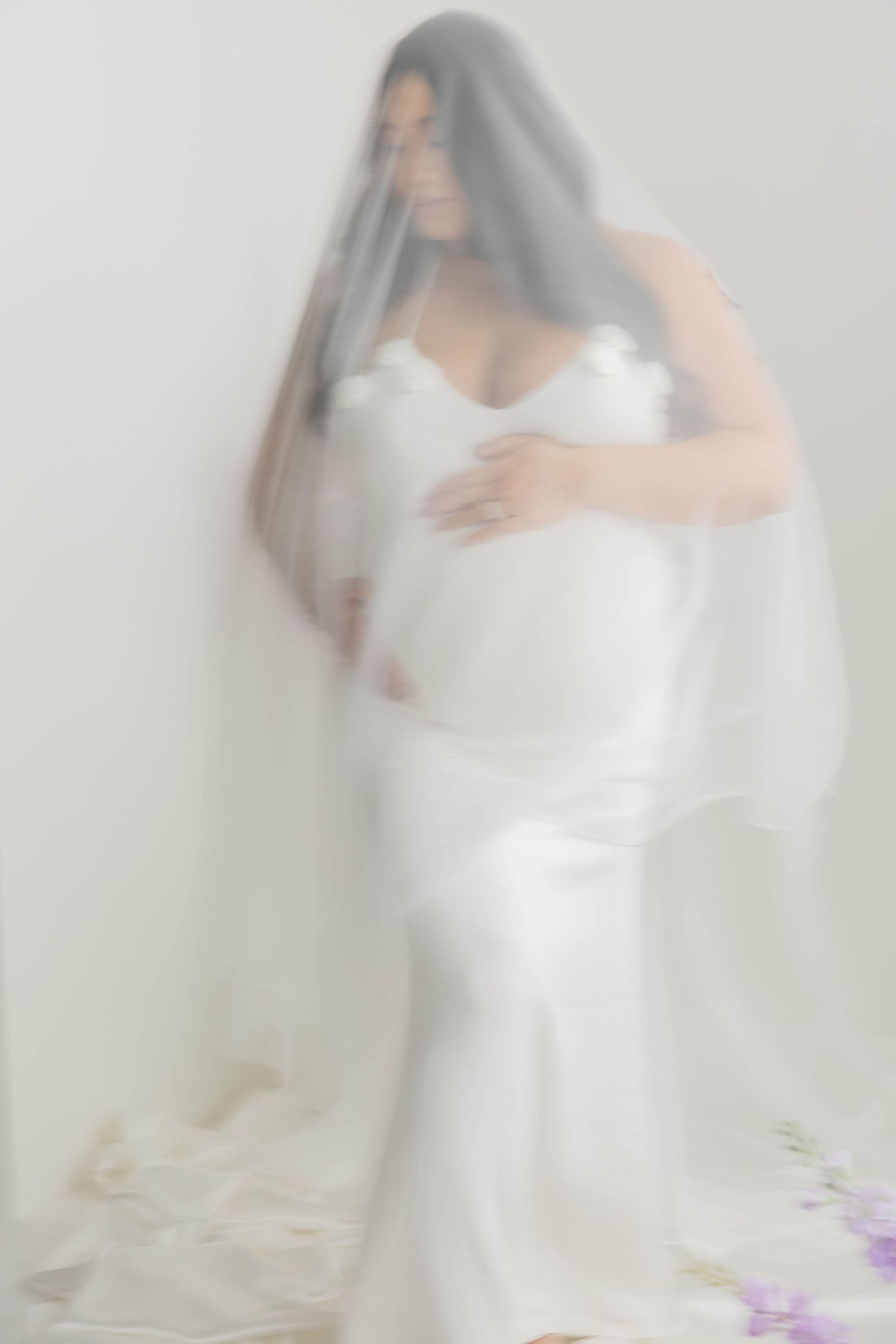 Ethereal maternity portrait with flowing white fabric and purple flowers creating a dreamy atmosphere.