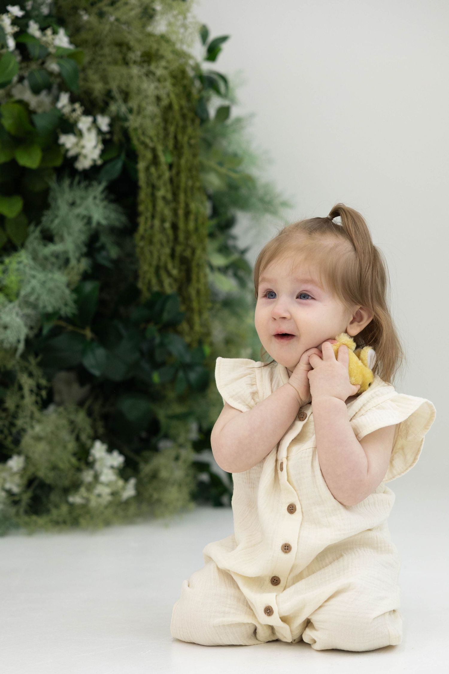 A toddler in a cream-colored ruffled romper sits on a white floor against a backdrop of lush green foliage.