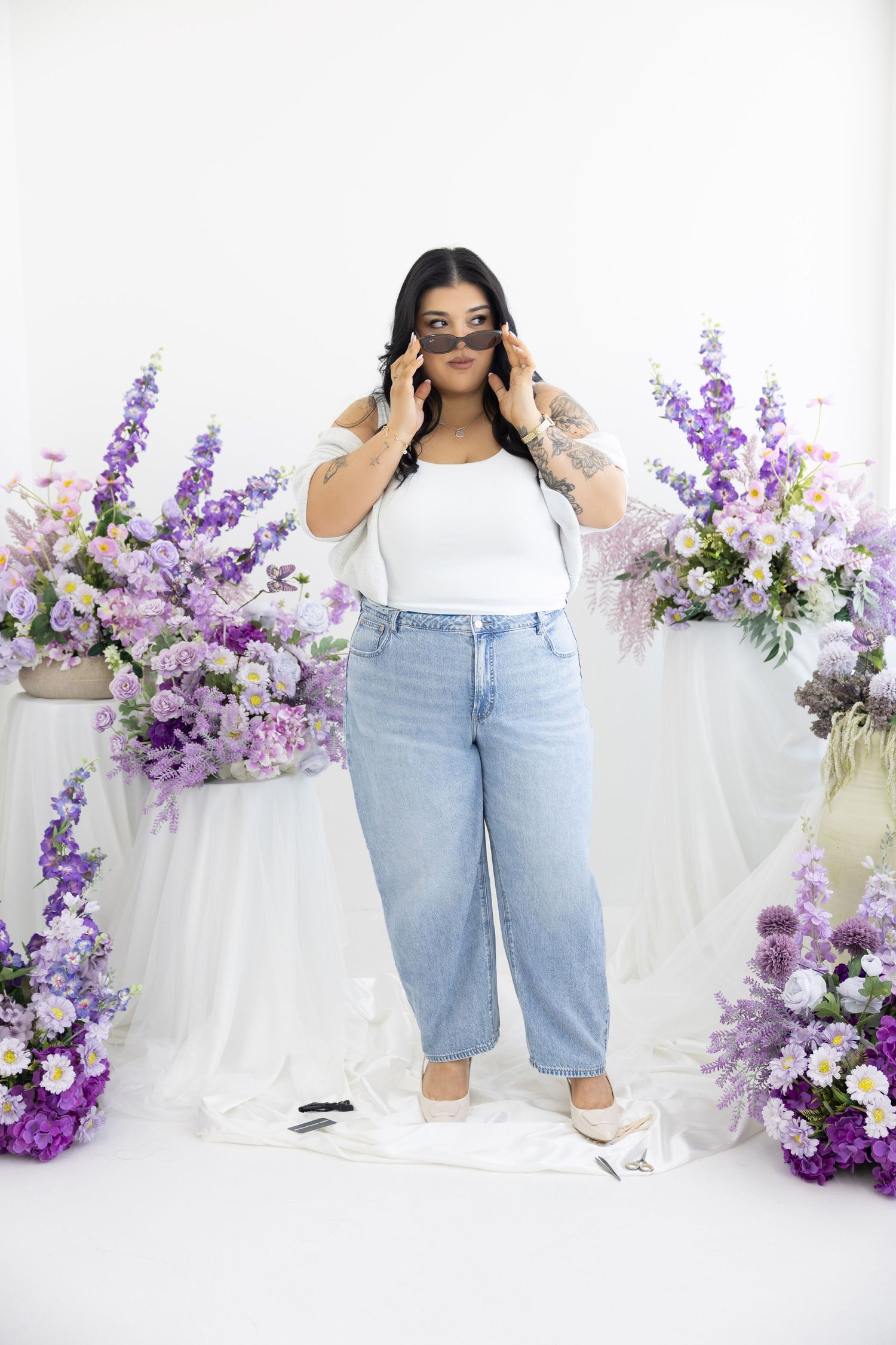 Woman in white top and light blue jeans posing with sunglasses surrounded by purple floral arrangements on white background.