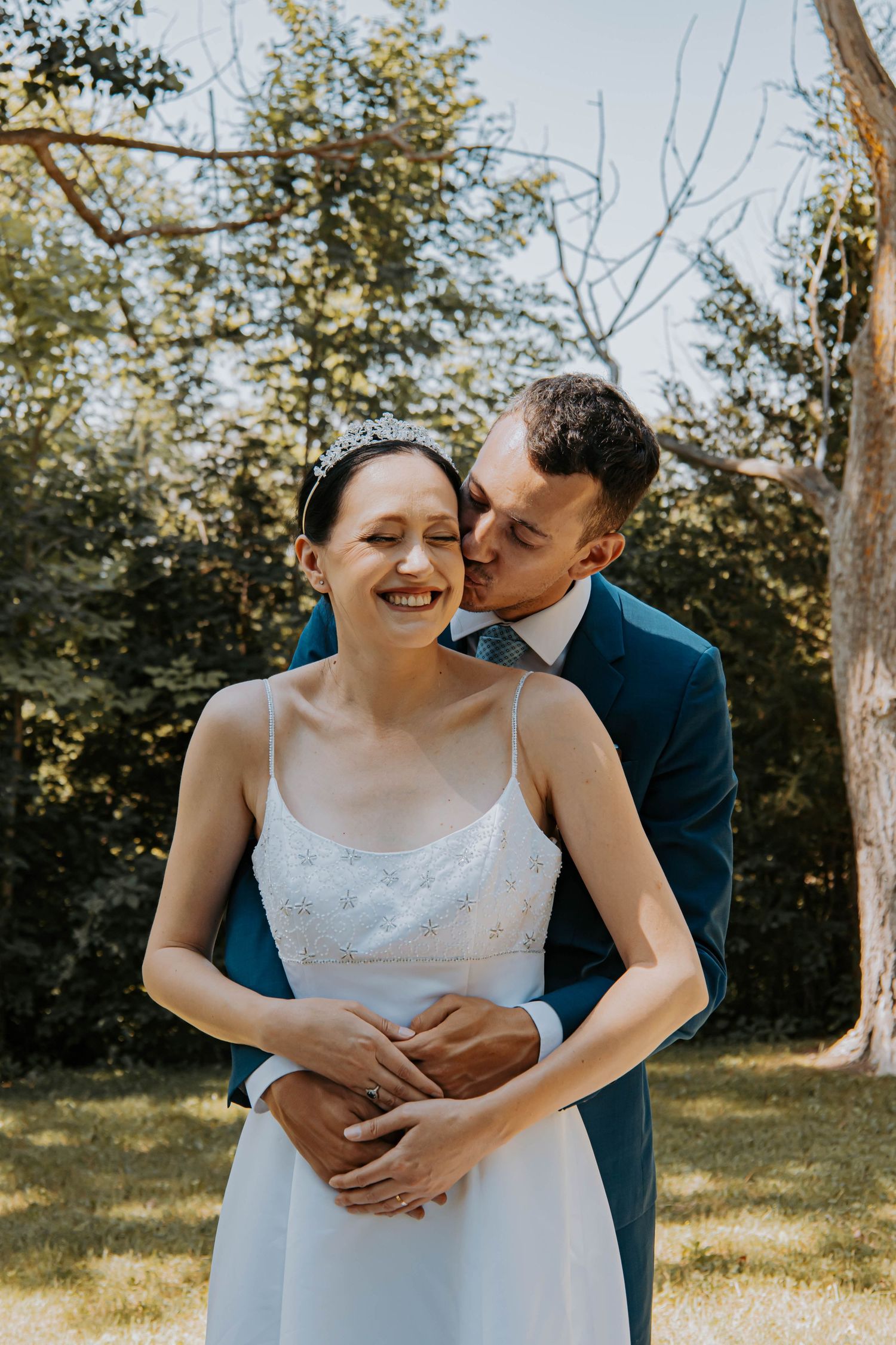 A tender moment between a couple in wedding attire beneath tall trees.