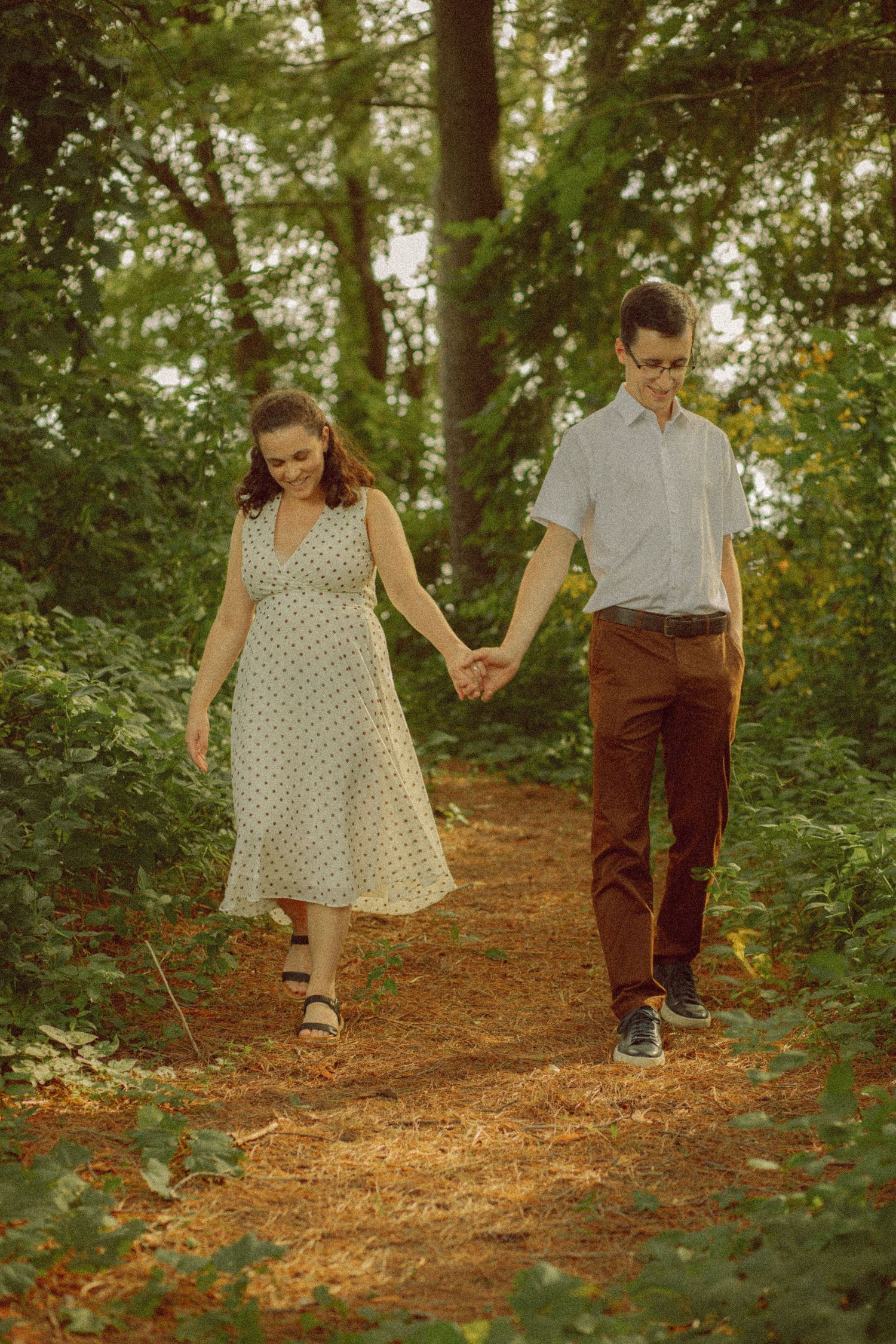 A couple holds hands while walking along a forest path, the woman in a polka dot dress and the man in brown pants.
