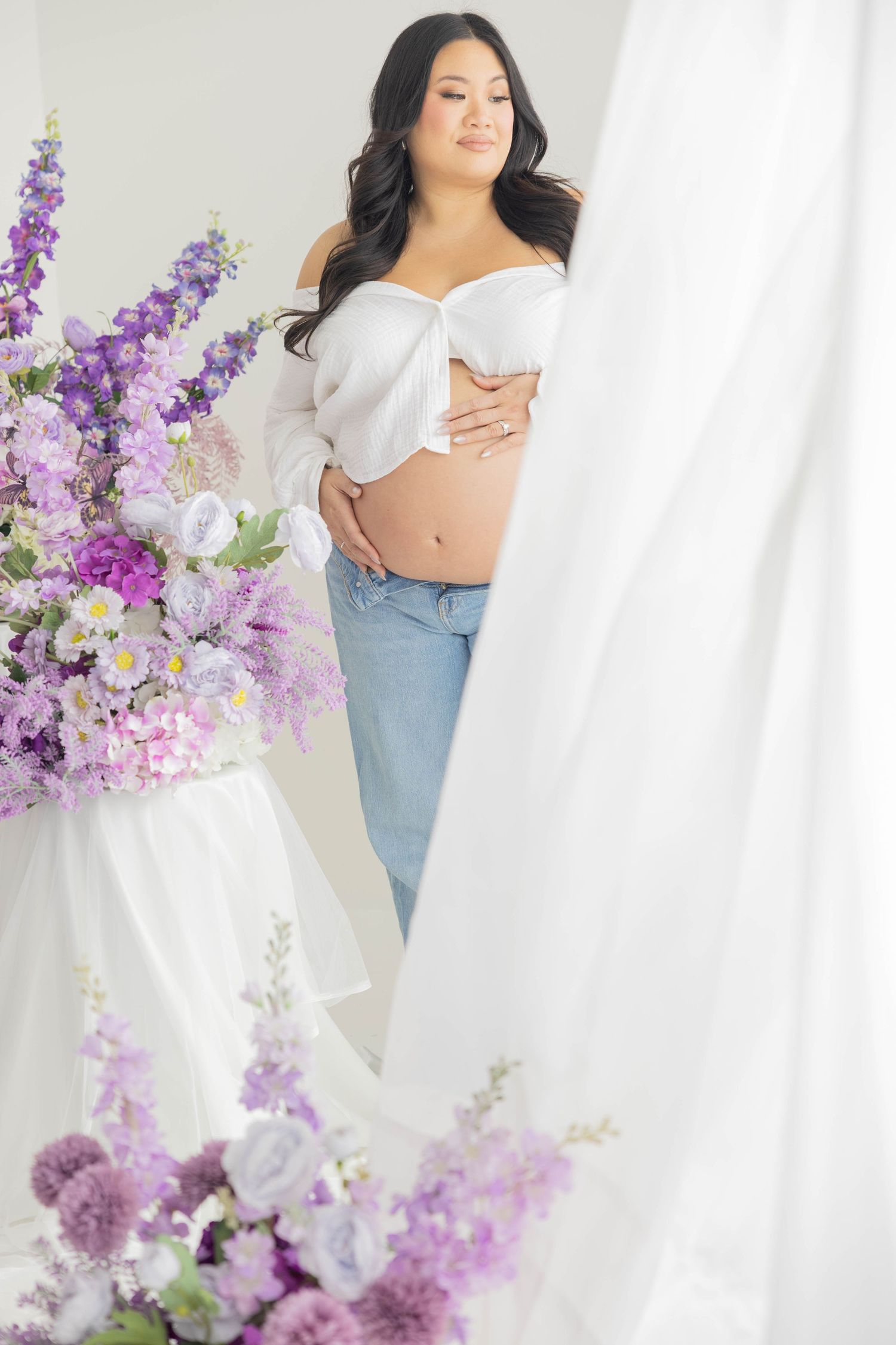 A woman in a white top and jeans poses elegantly with purple lilac flowers against a white backdrop.
