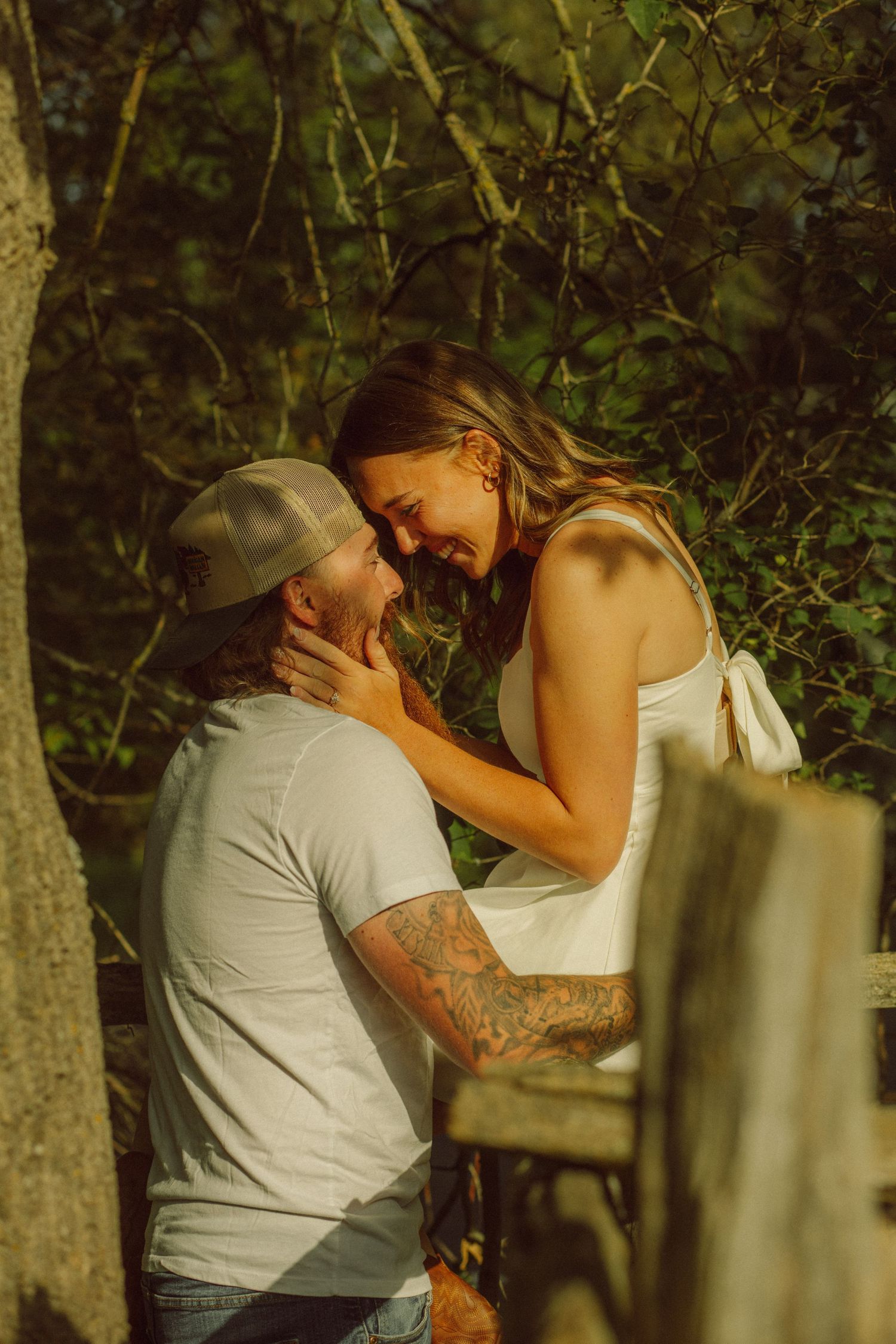 Tattooed man in cap and woman in white dress share a romantic laugh outdoors in golden sunlight near trees.