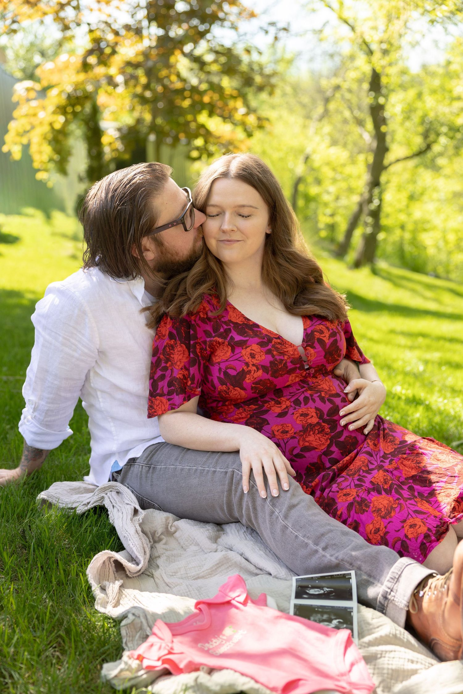 A couple sits on a blanket in a sunny park, man kissing pregnant woman's cheek as she smiles peacefully.