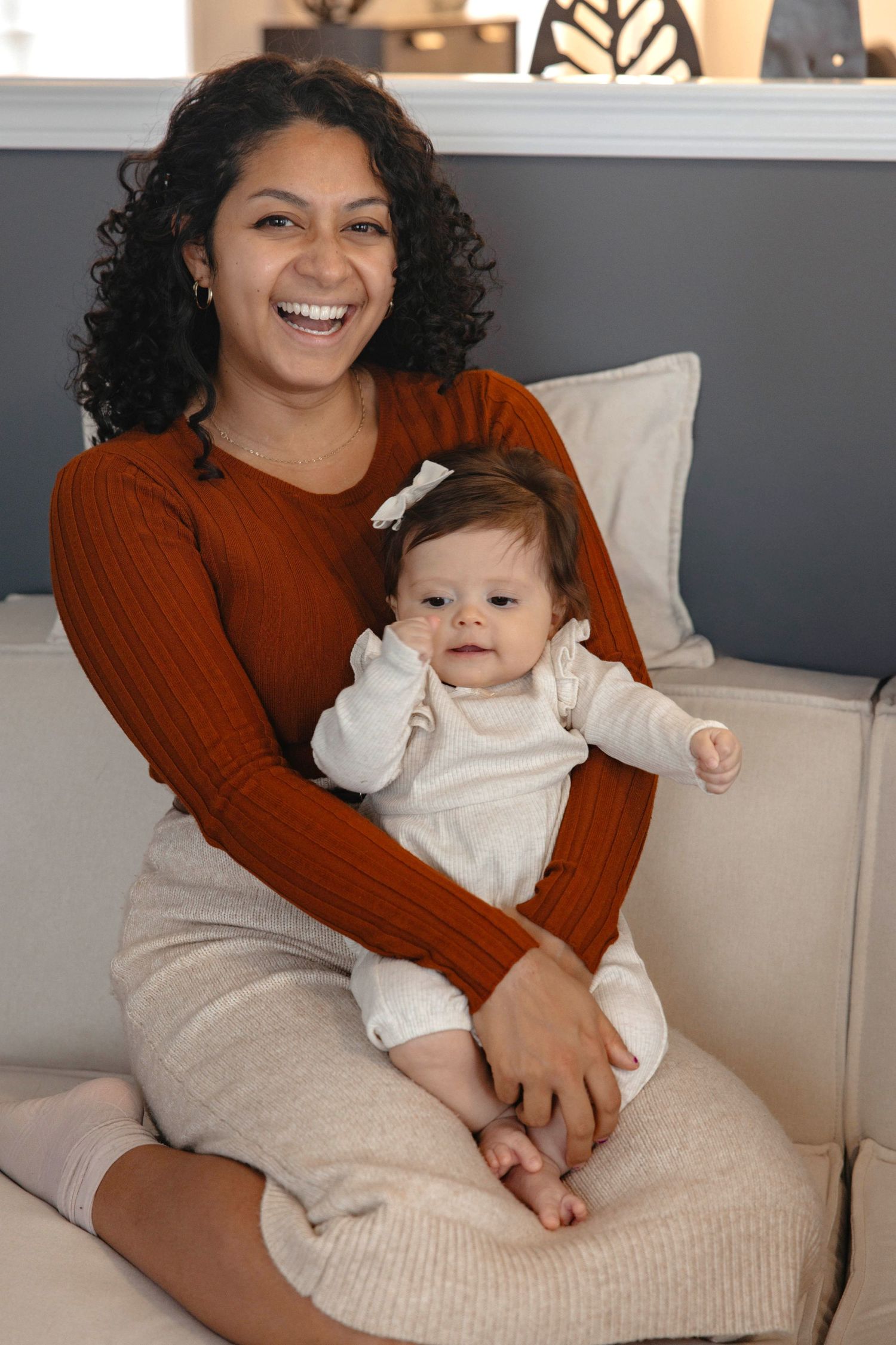 Mother and baby share joyful moments together on a white couch with grey walls in background.
