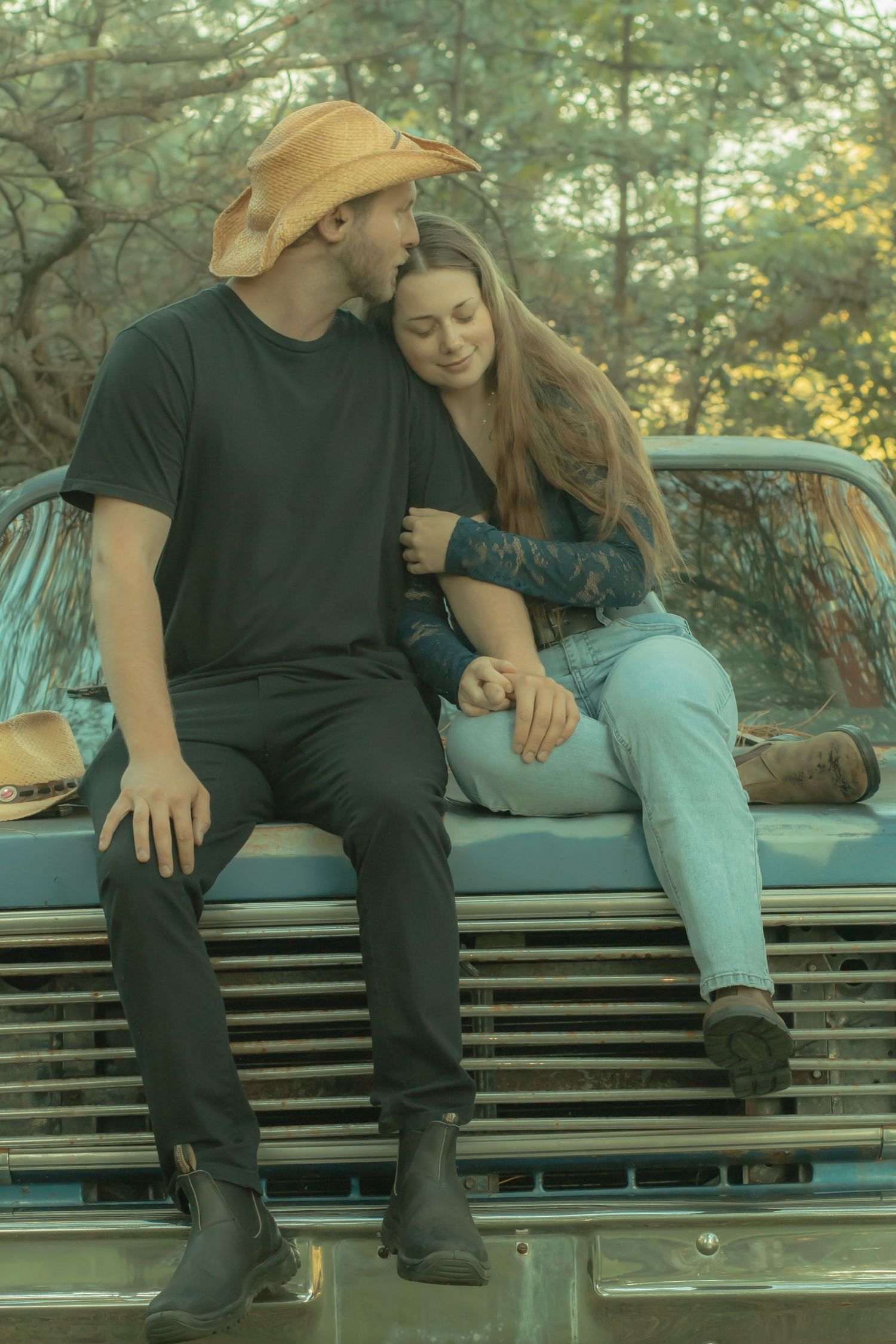 Romantic couple sitting on vintage truck hood, man in cowboy hat kissing woman's head in forest setting.