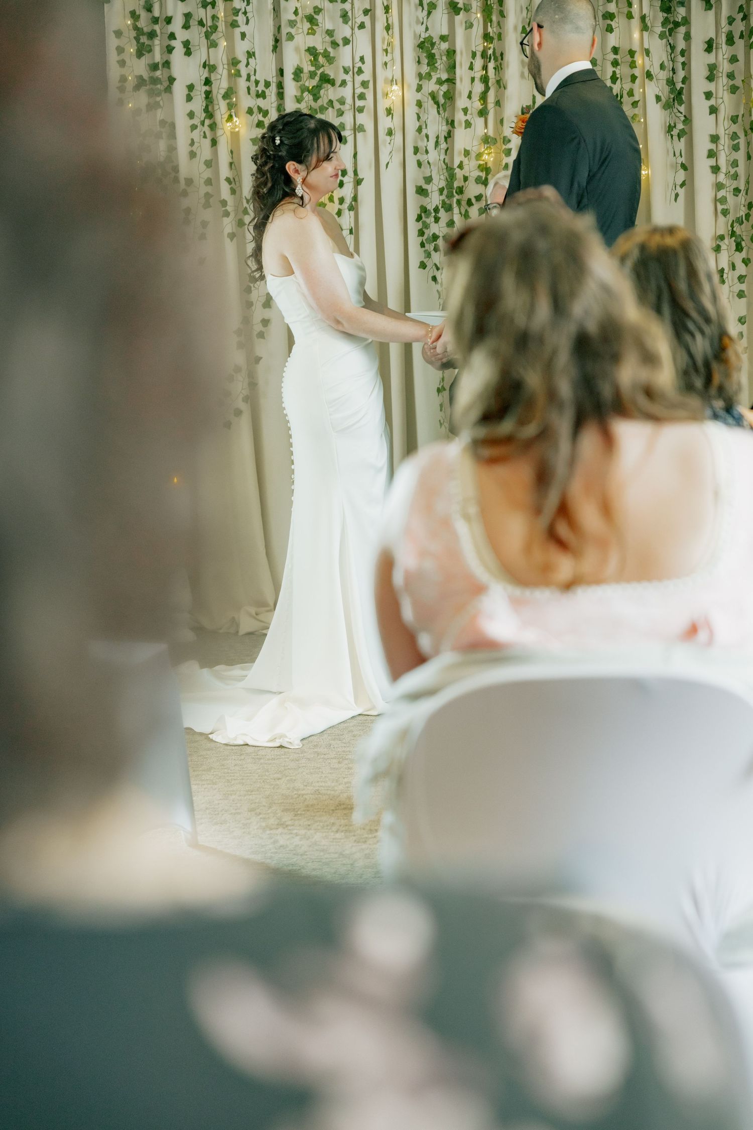 Bride in white gown holds hands with groom at altar decorated with greenery while guests watch the ceremony.