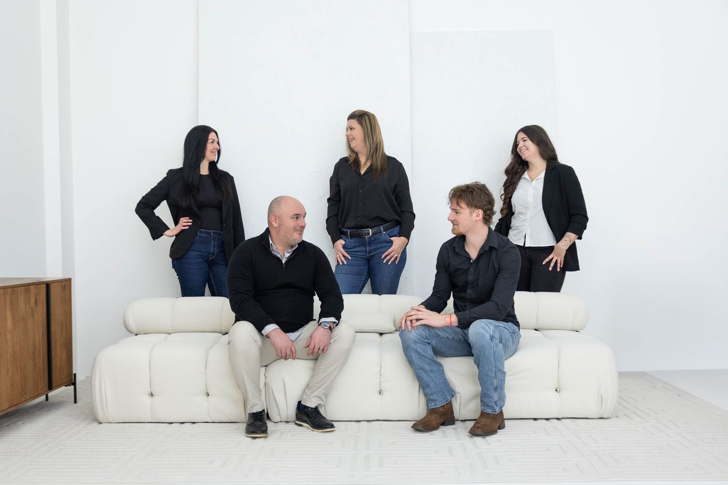 Five business professionals in casual attire sit and stand around a white couch in a minimalist studio setting.
