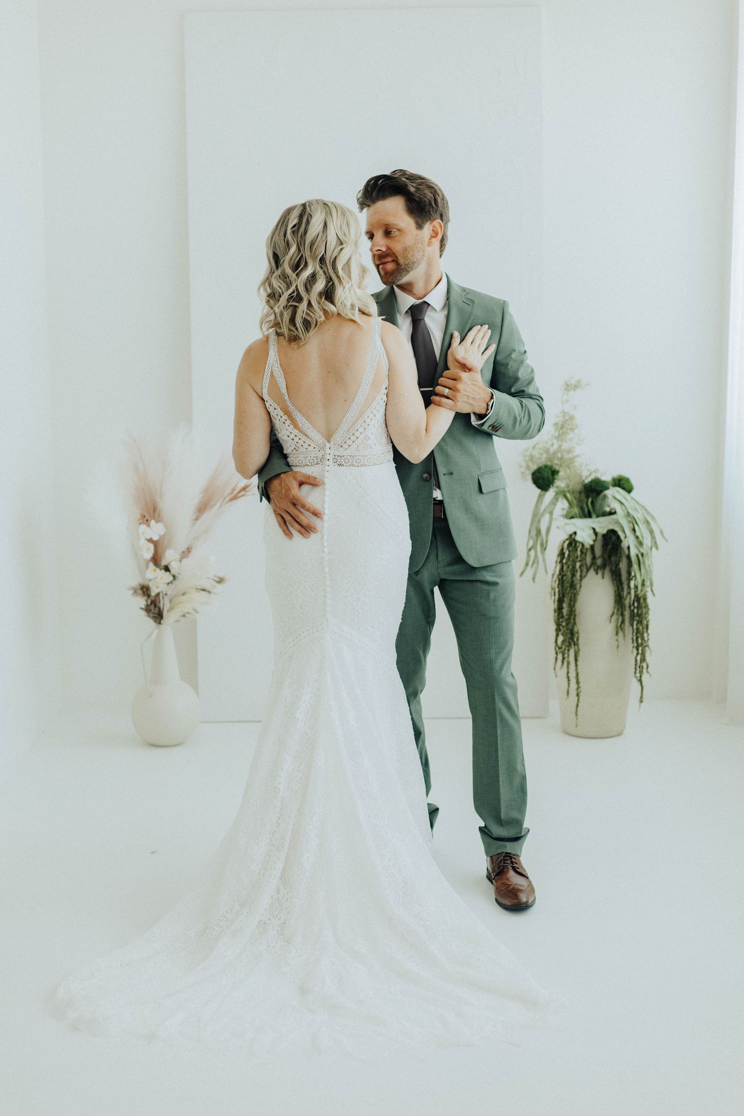 A couple in wedding attire dancing together in an elegant white studio space with potted plants in the background.