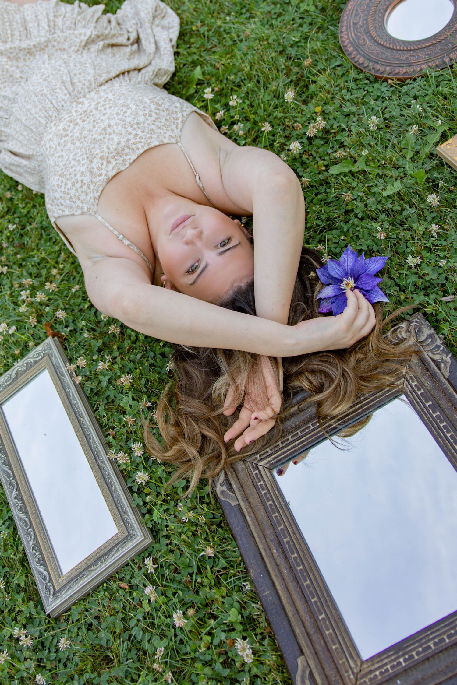A person lies on grass beside an ornate picture frame while holding a purple flower in an artistic composition.