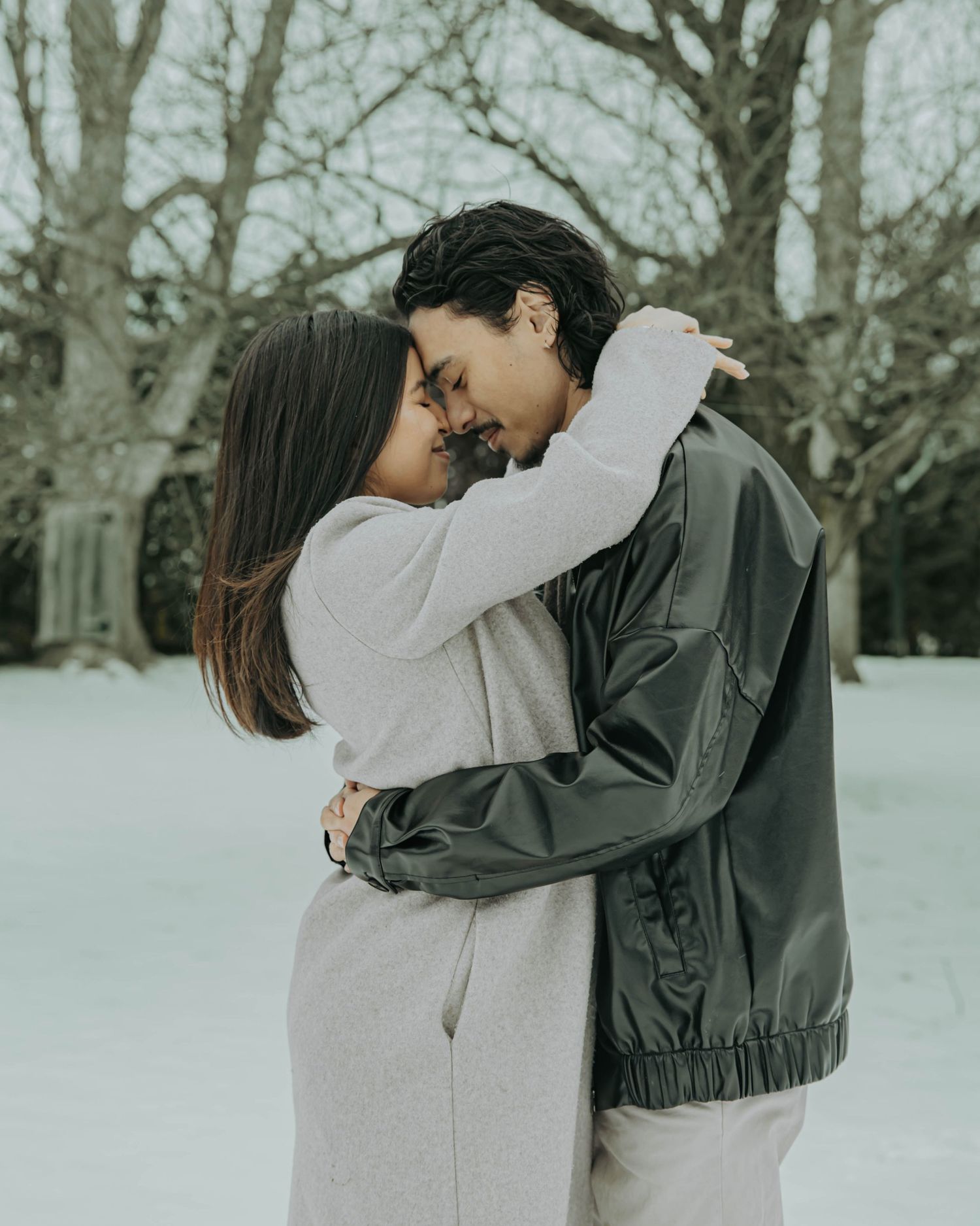 A couple embraces in a romantic moment on a snowy winter day, with bare trees in the background.