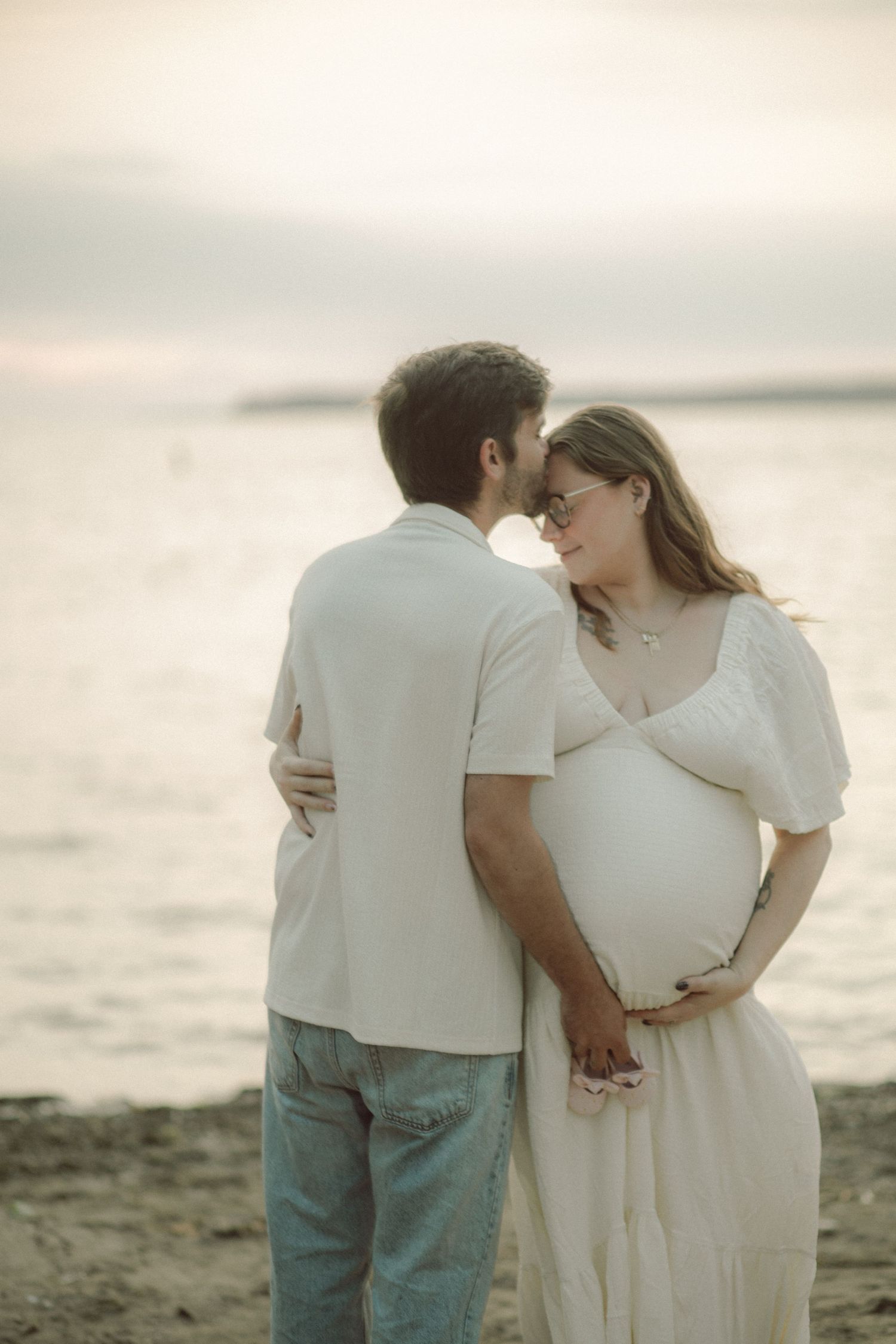 Pregnant couple sharing a tender moment on a sandy beach at sunset, woman in white dress cradling her baby bump.