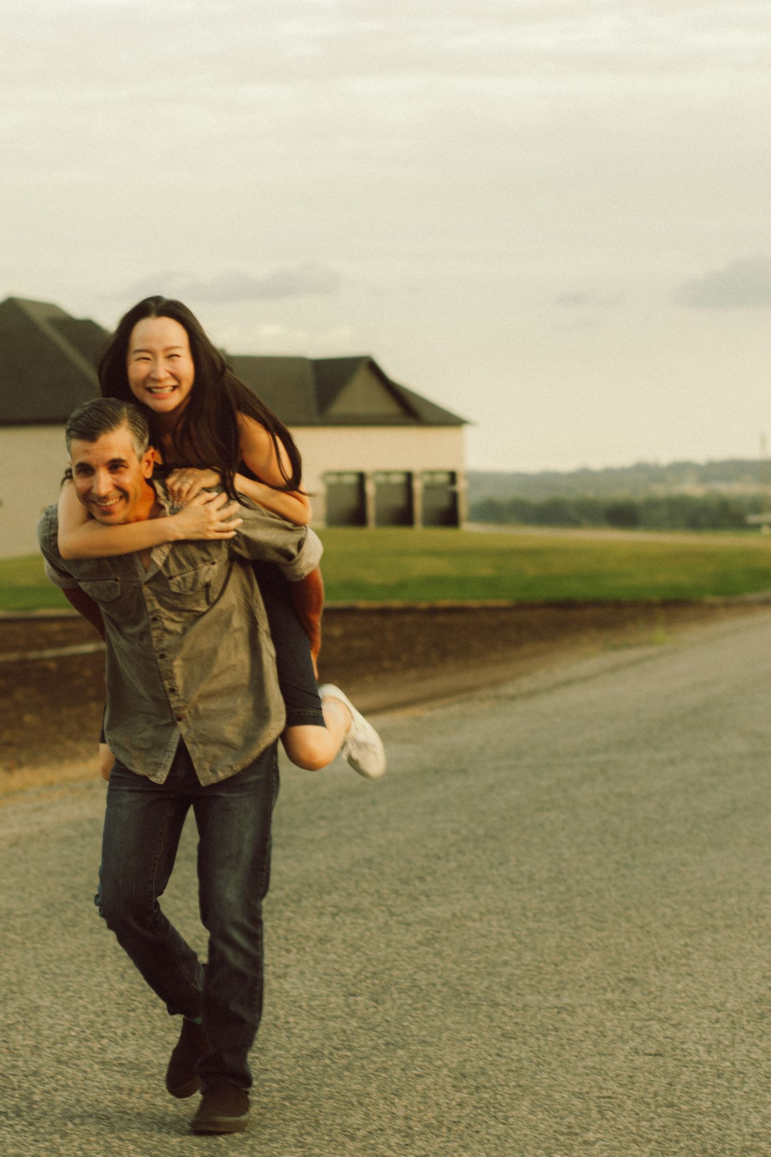 A happy couple shares a piggyback ride on a rural road, smiling joyfully with green fields and a house in the background.