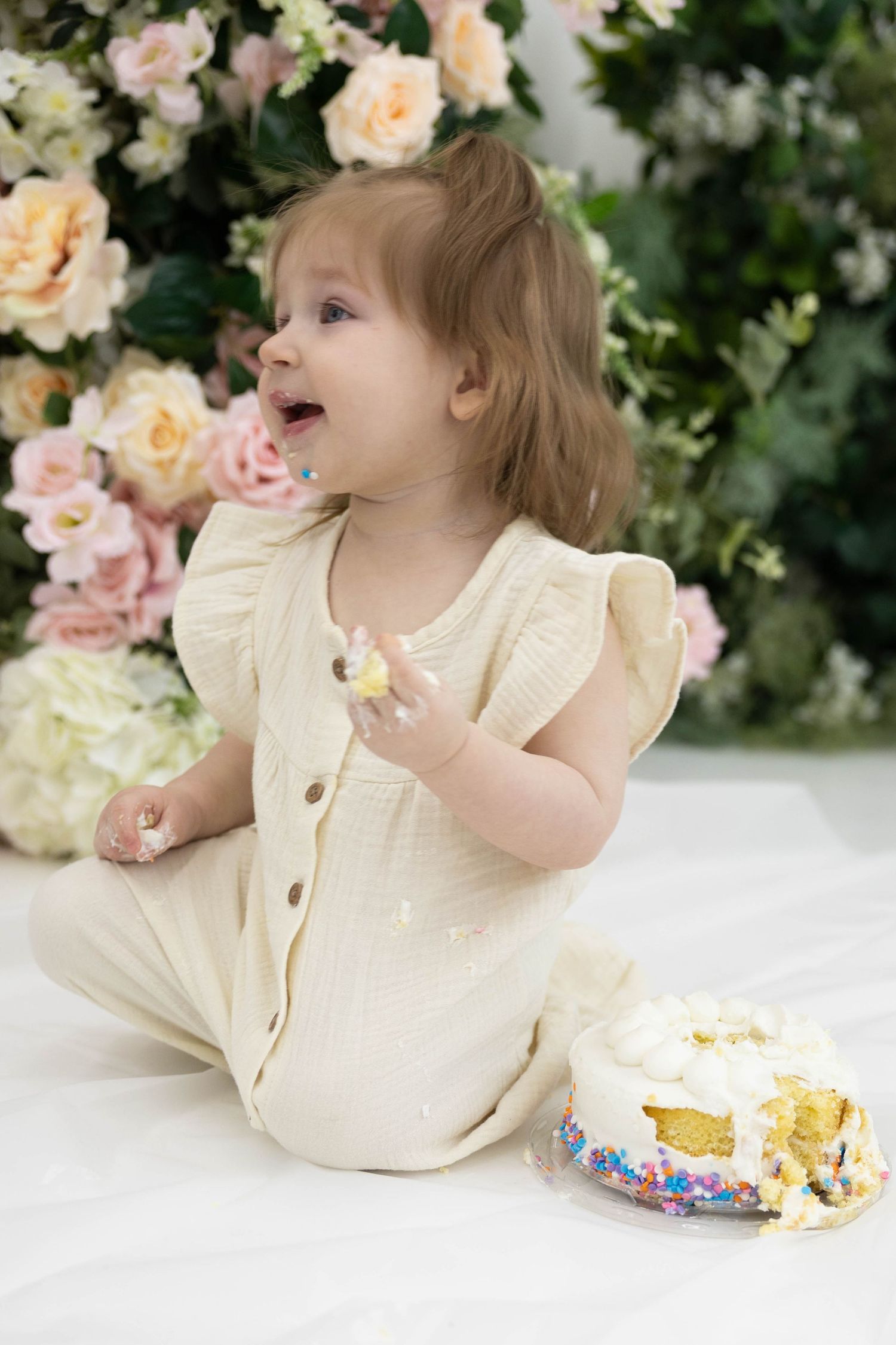 A young child in a light-colored outfit smiles joyfully next to a birthday cake with pink roses in the background.
