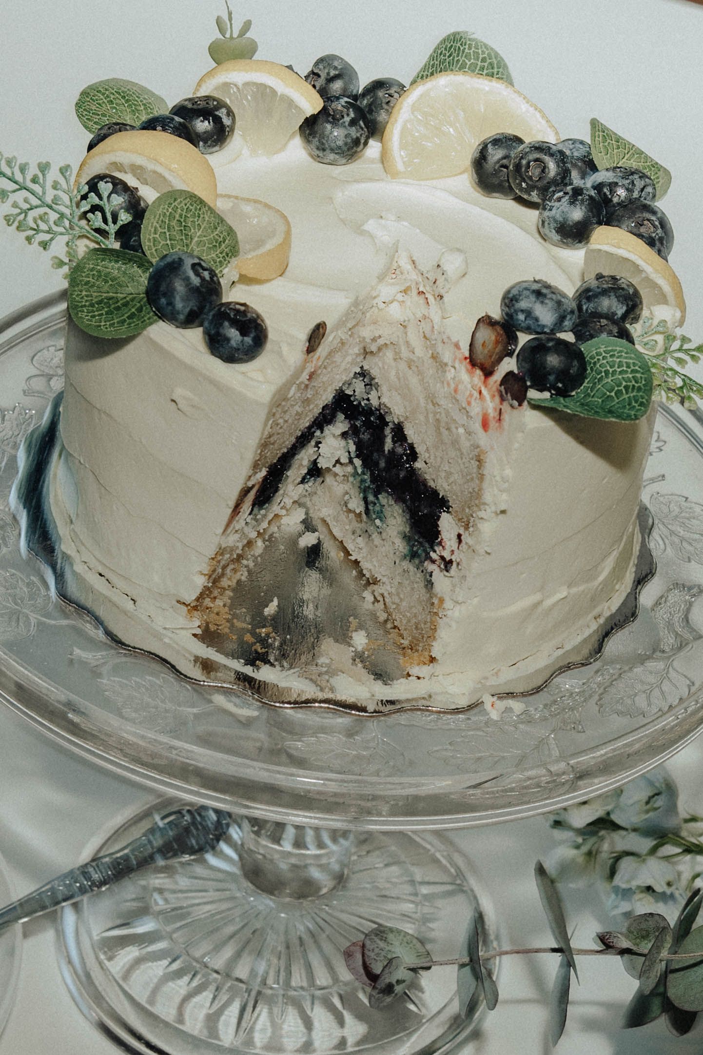 A white wedding cake decorated with fresh blueberries and mint leaves on a crystal cake stand.
