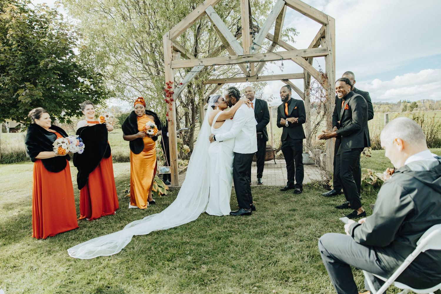 Outdoor wedding ceremony setup with wooden arch structure and guests seated on chairs in a natural setting.