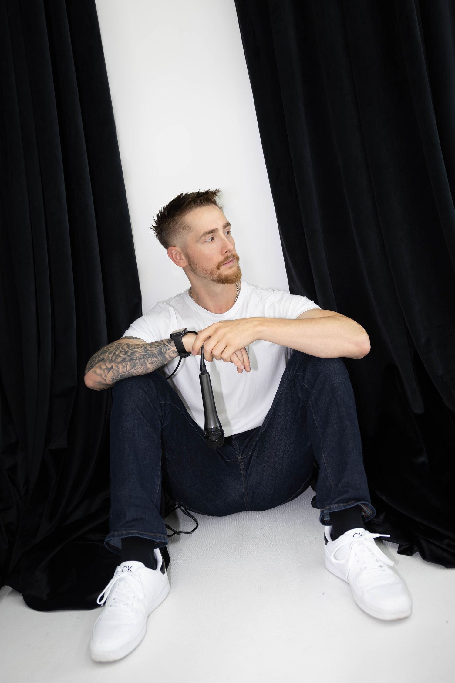 Person in white shirt and dark pants sits casually against black backdrop wearing white sneakers.