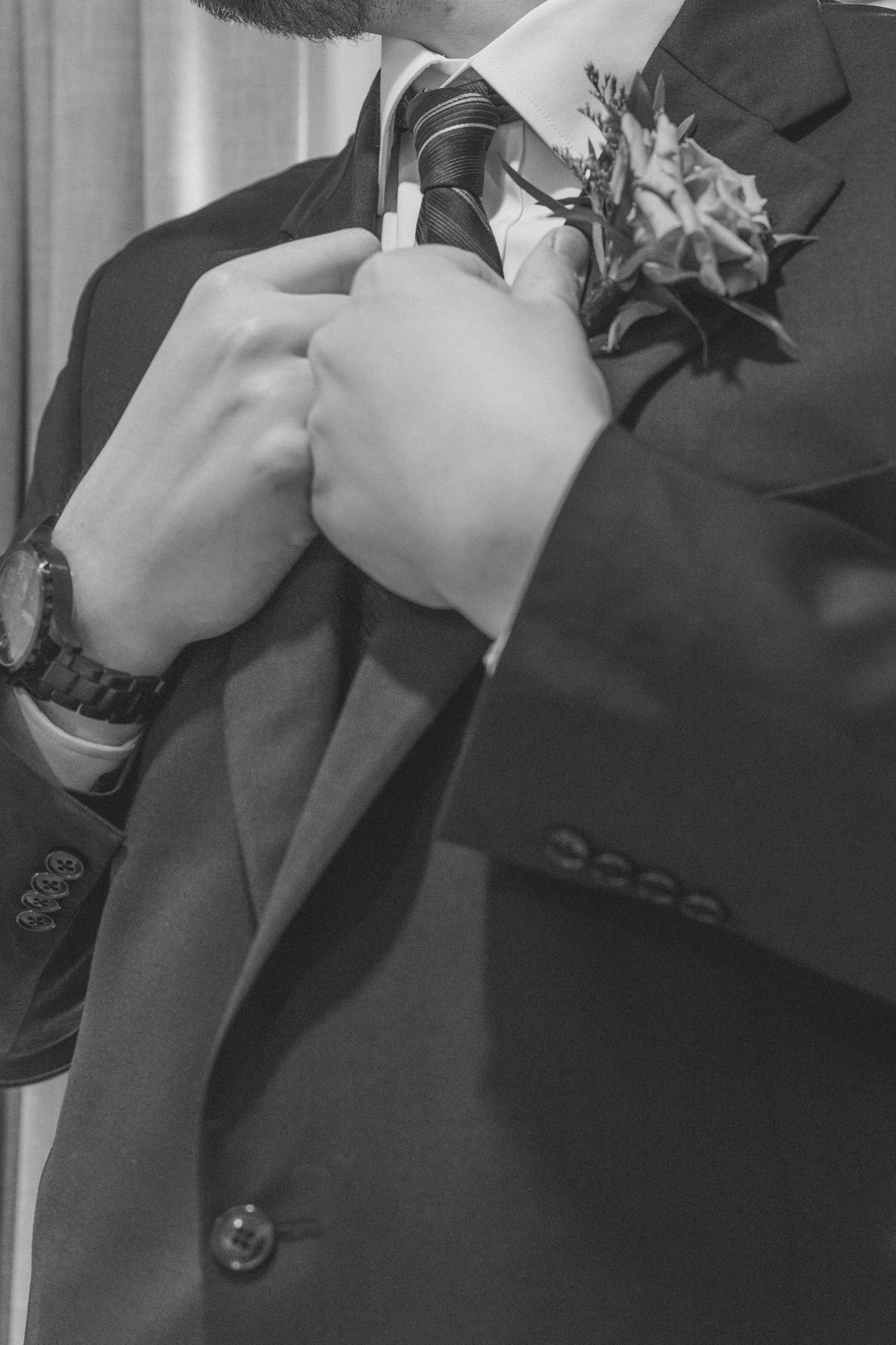 Black and white photo of a groom adjusting his tie, wearing a suit with a floral boutonniere and a watch.