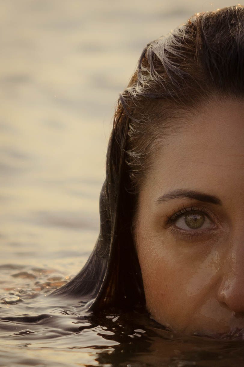 Close up shot of a person partially submerged in water during sunset with sun reflecting in their eye.