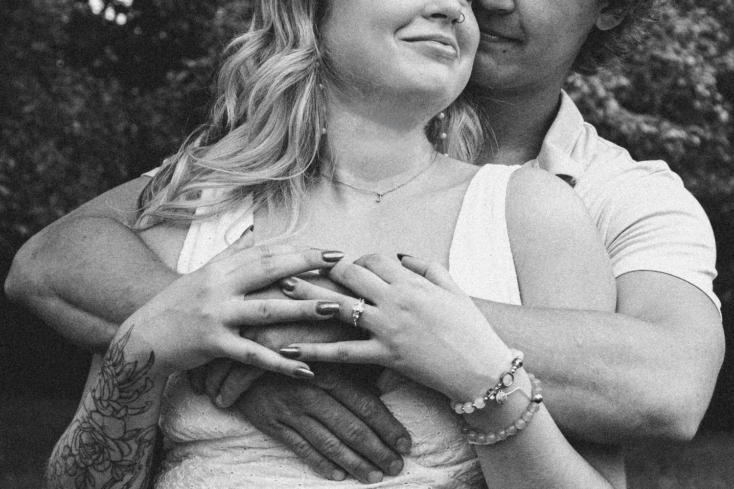 A close-up black and white engagement photo showing clasped hands with a bracelet and tattoo detail.