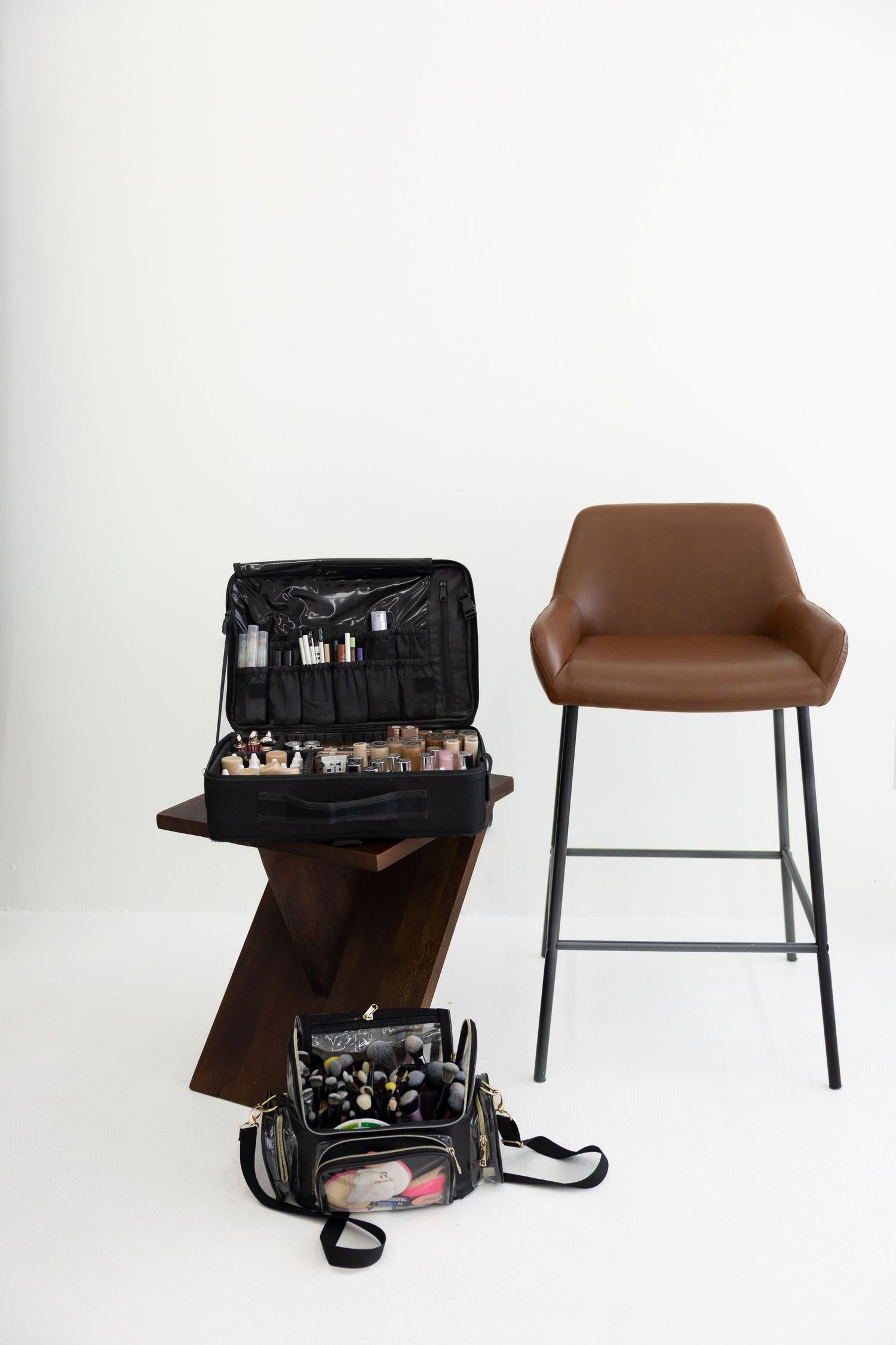 An open professional makeup kit on a wooden stand beside a brown leather bar stool against a white wall.