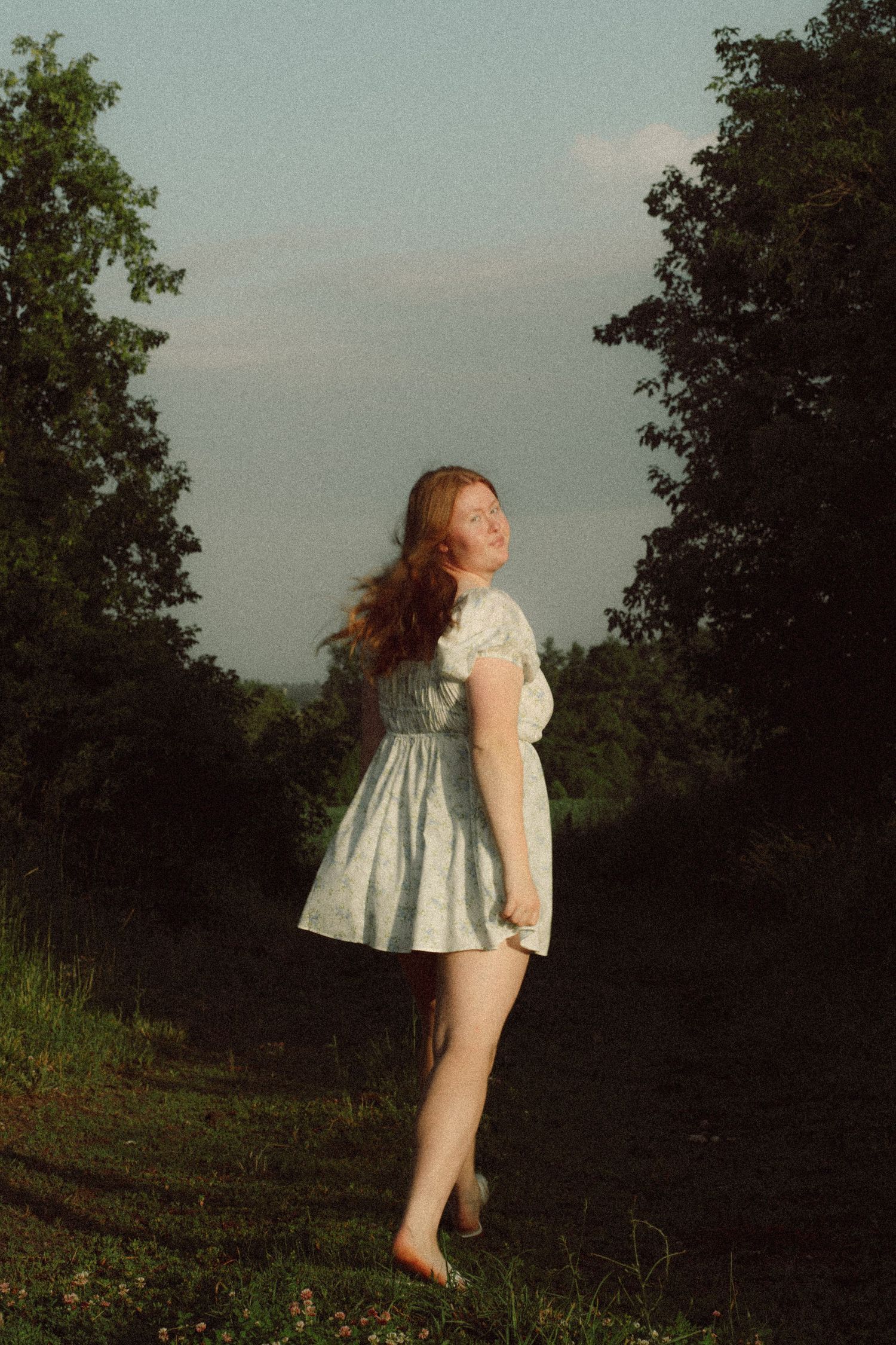 Young redhead woman in white mini dress standing barefoot on rural path between trees at sunset.
