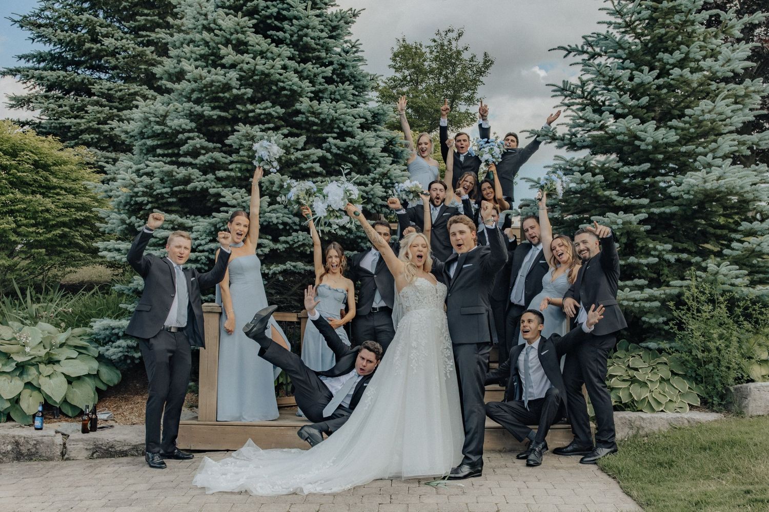 Wedding party celebrating outdoors with bride and groom, bridesmaids in blue, groomsmen in black, pine trees background.