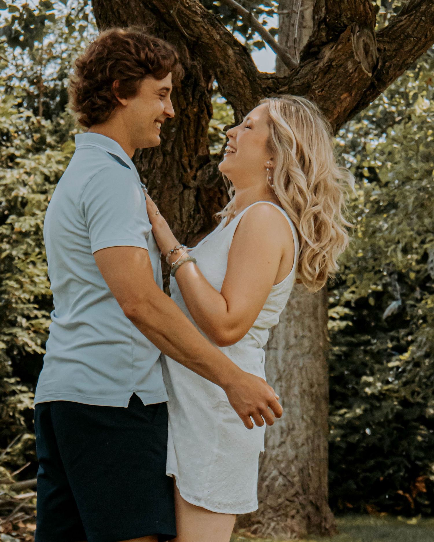 Romantic couple sharing tender moments in a natural outdoor setting under a large tree in summer lighting.
