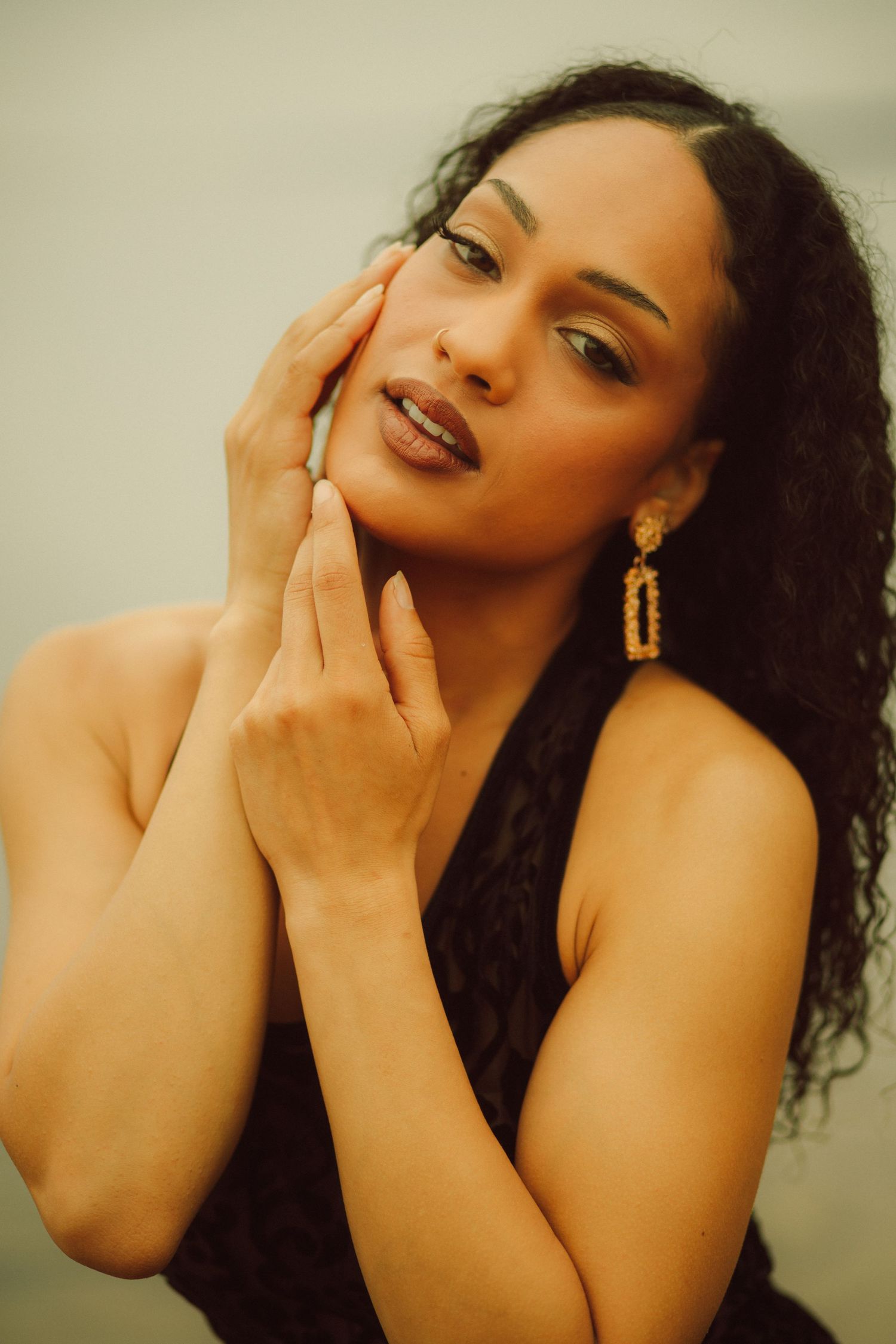 A woman with curly hair wearing gold earrings and black top poses gracefully with hands near her face.